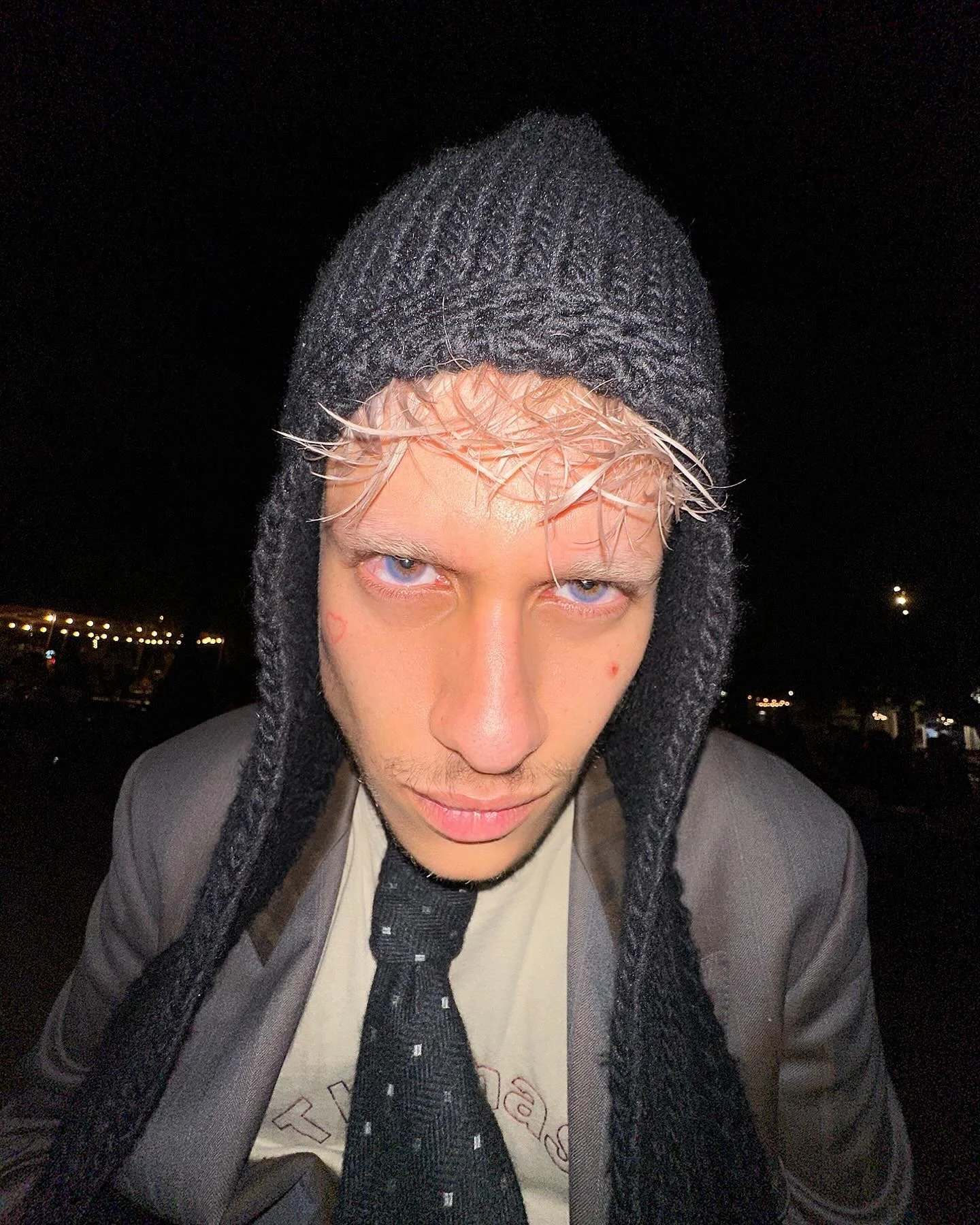 Jeune homme portant un bonnet en laine grise, une chemise beige, une cravate noire, et un blazer, regardant directement la caméra sous un ciel nocturne avec des lumières en arrière-plan.