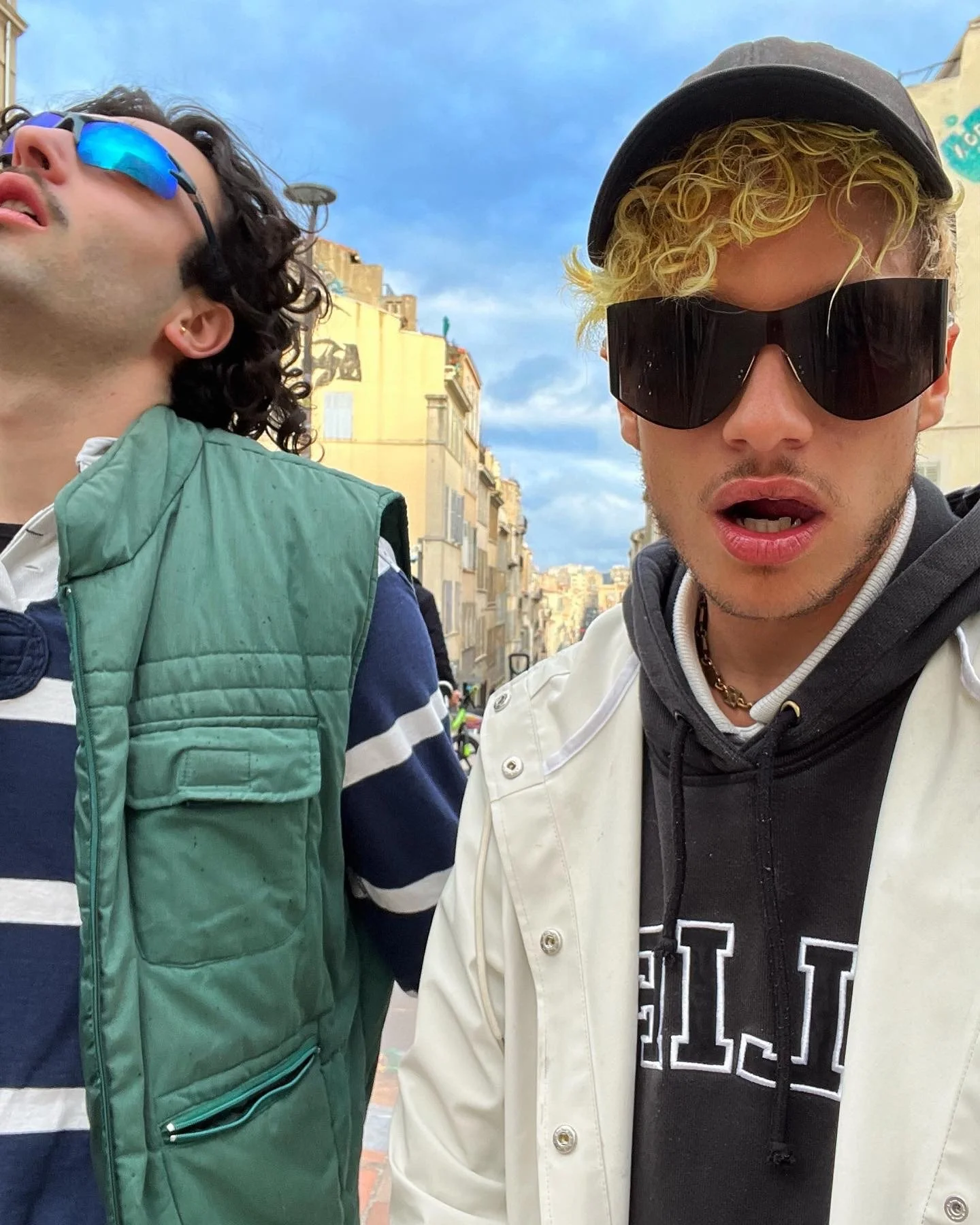 Deux jeunes hommes portant des lunettes de soleil dans une rue urbaine, avec des bâtiments en arrière-plan et un ciel bleu avec quelques nuages