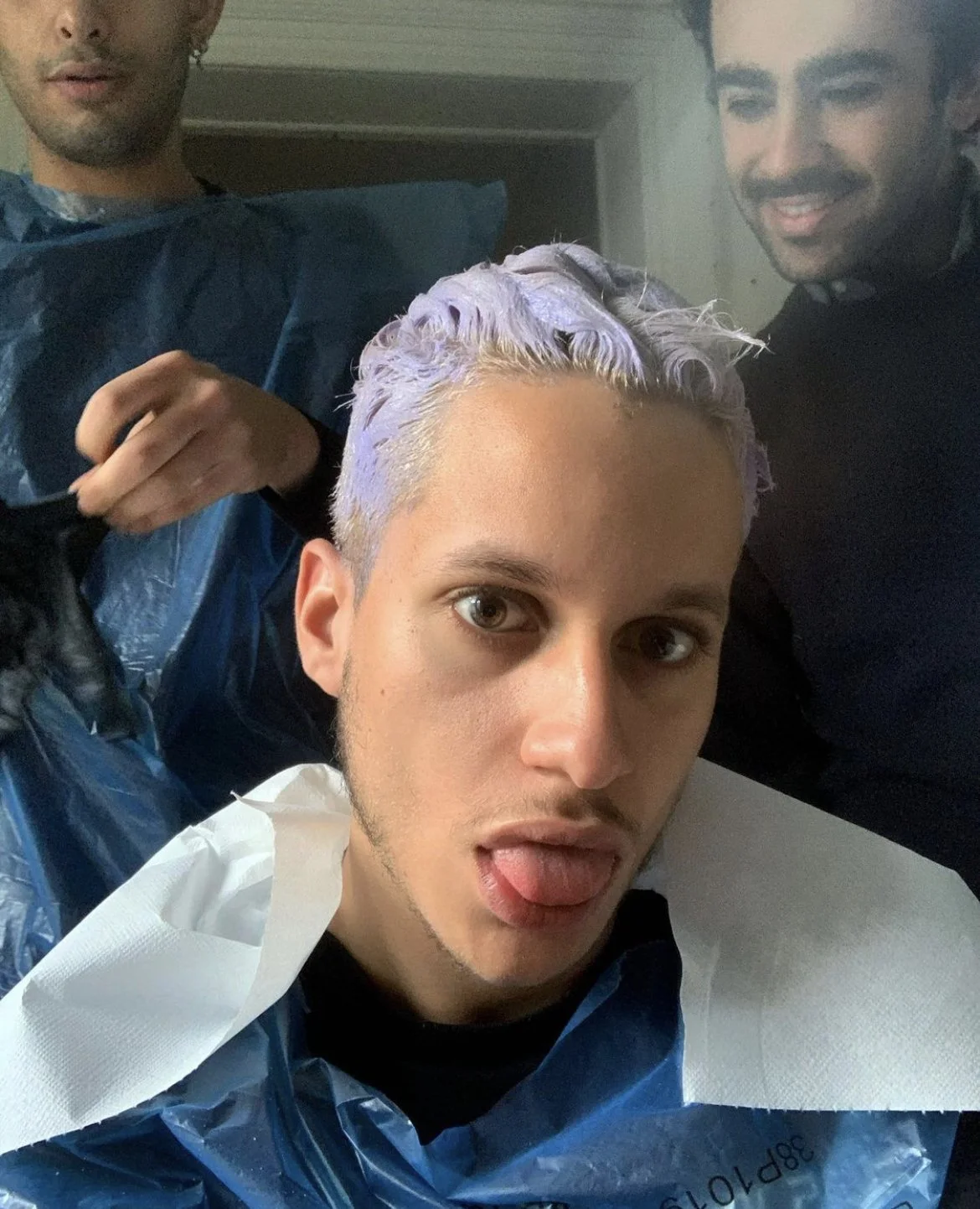 Un jeune homme se fait teindre les cheveux en violet par deux assistants dans un salon de coiffure, faisant une expression amusante avec la langue sortie.
