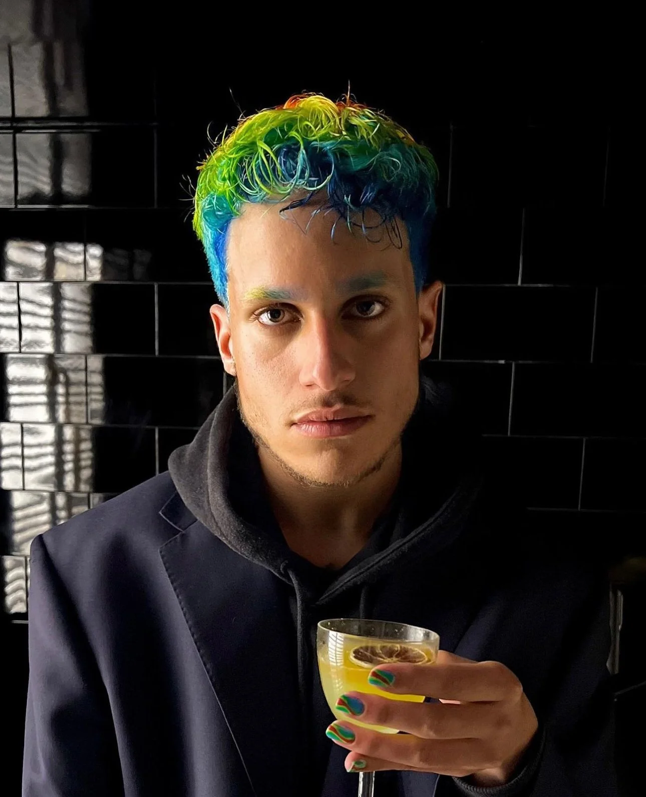 Un jeune homme avec des cheveux colorés en arc-en-ciel, tenant un verre de cocktail jaune, enroulé dans un décor sombre.