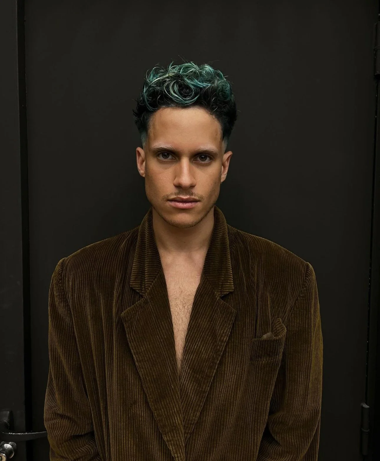 Jeune homme avec cheveux bouclés teints en vert et noir, portant une veste en velours marron, posant devant un mur noir.