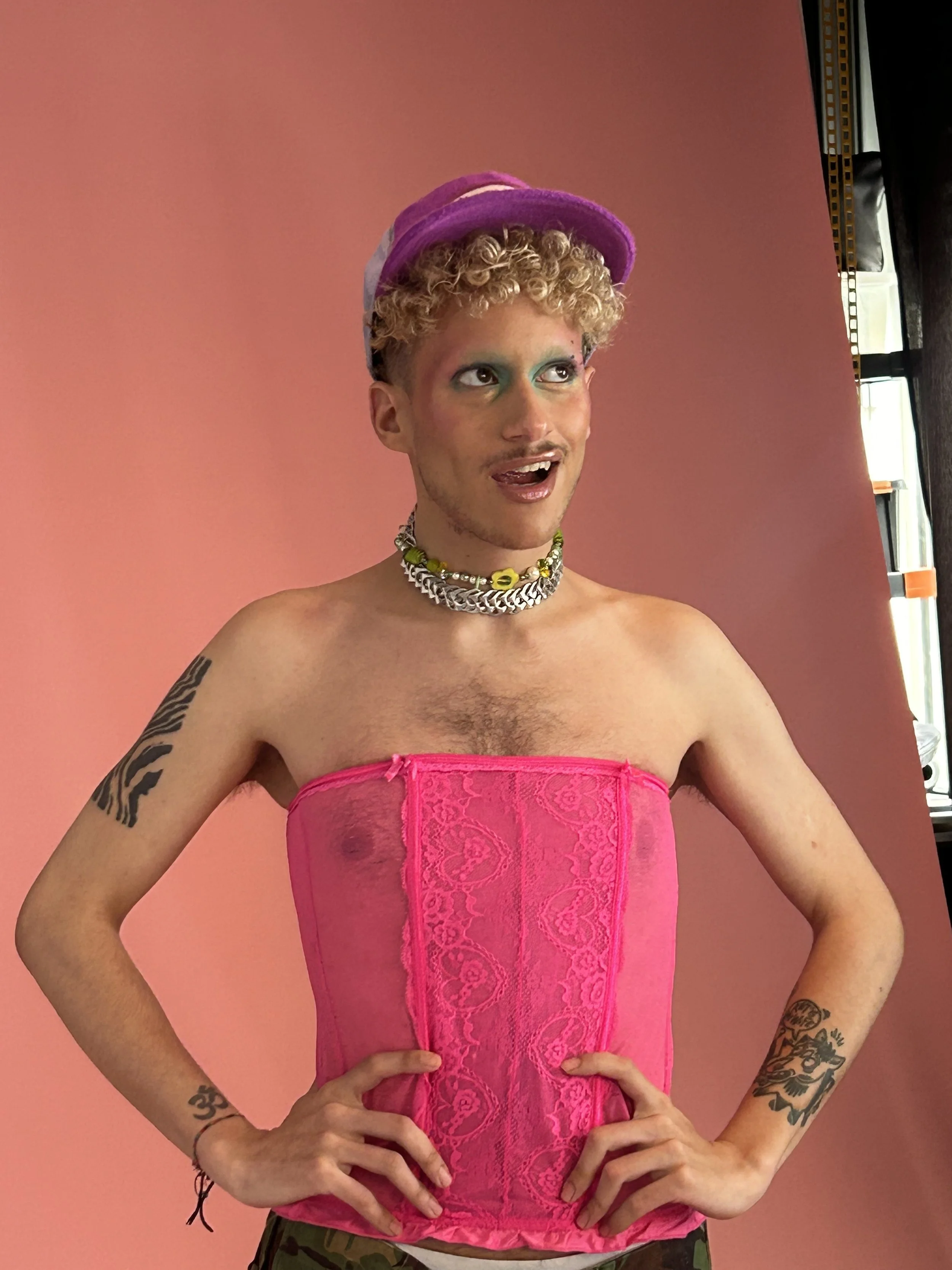 Une personne avec un maquillage coloré, portant un bandeau violet, un collier coloré, un corset rose en dentelle, et affichant plusieurs tatouages, se tient contre un mur rose.