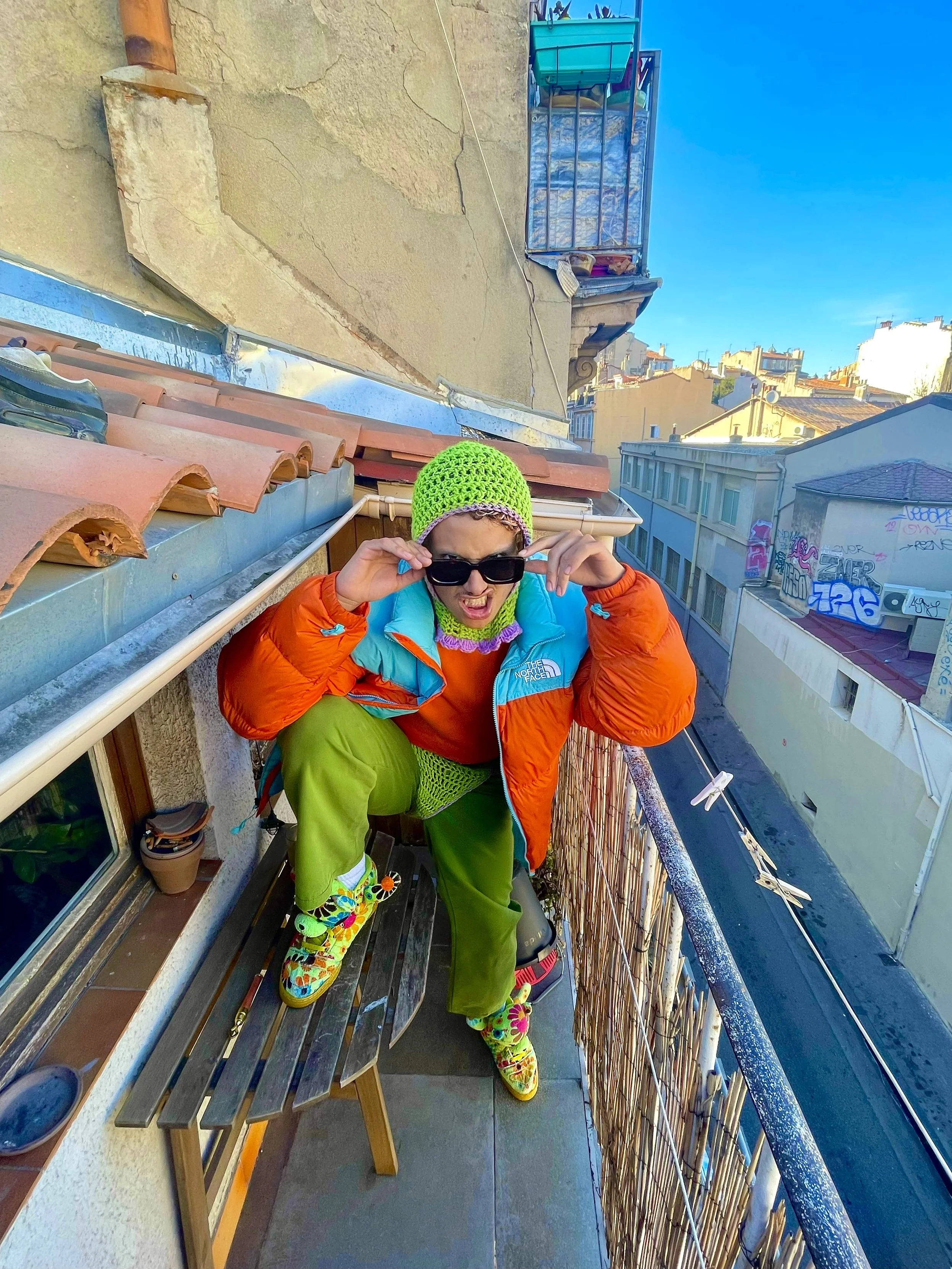Une personne en vêtements colorés debout sur un balcon, portant des lunettes de soleil, un bonnet vert, une veste orange et des chaussures multicolores.