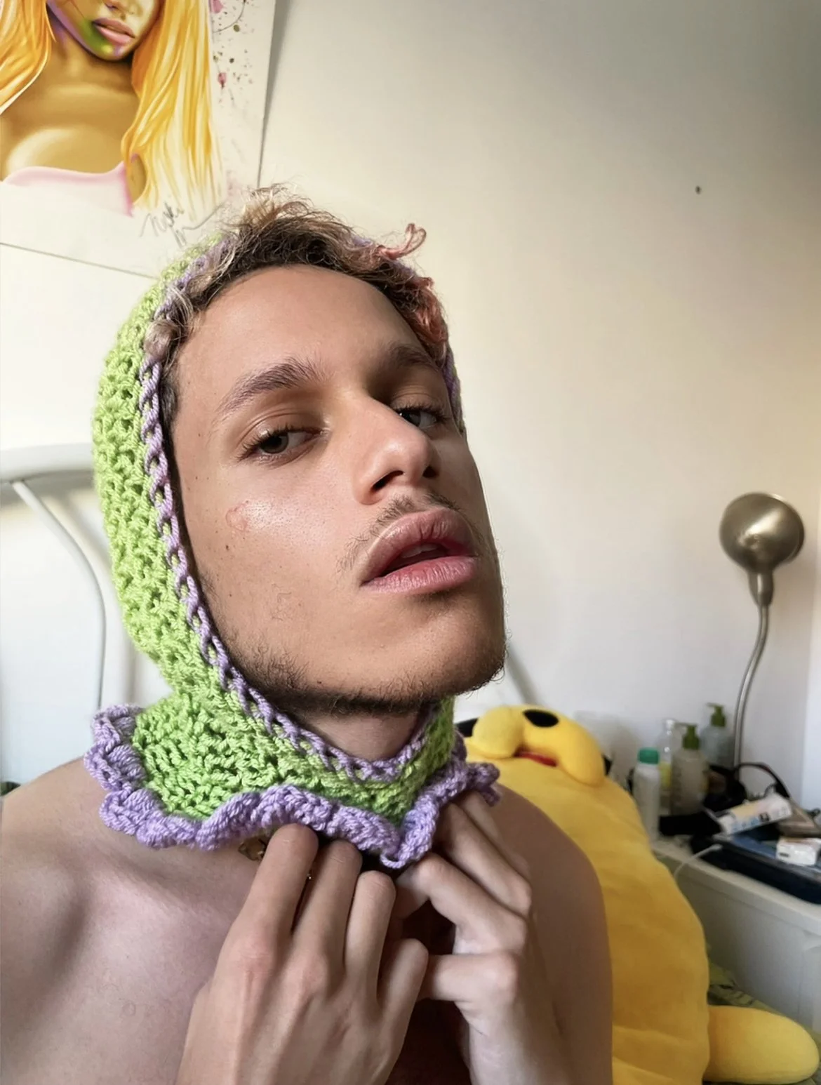 Jeune homme portant un bonnet en crochet vert et violet, posant dans une chambre avec un lit jaune et des objets de toilette sur une table de chevet.