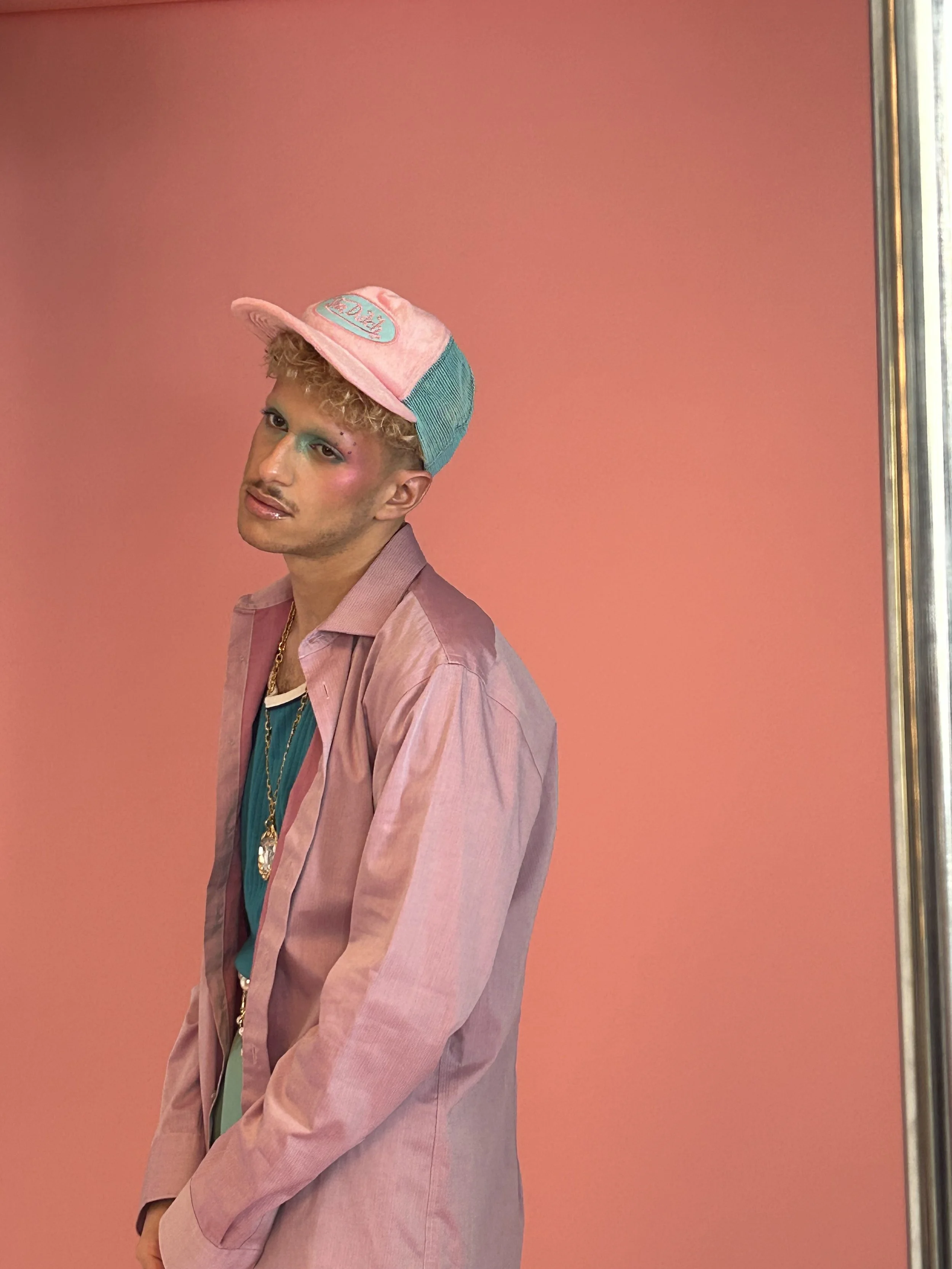 Jeune homme portant un chapeau de baseball rose et vert, maquillage coloré, veste rose, t-shirt bleu, pendentifs en or, posant devant un mur rose.