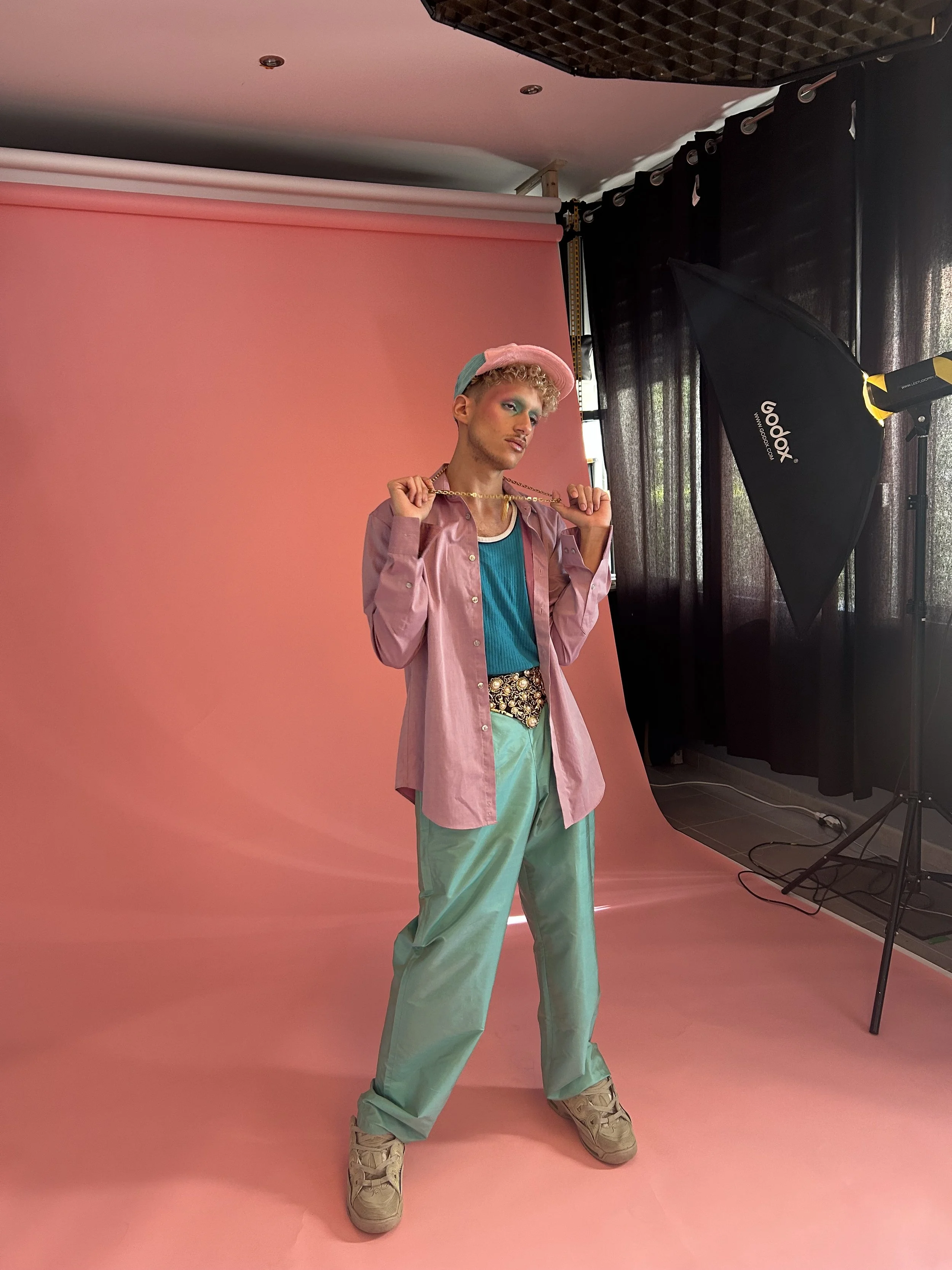 jeune homme posant avec une chaîne dorée, coiffé d'une casquette rose et portant une tenue colorée, dans un studio avec fond rose et équipement de photographie