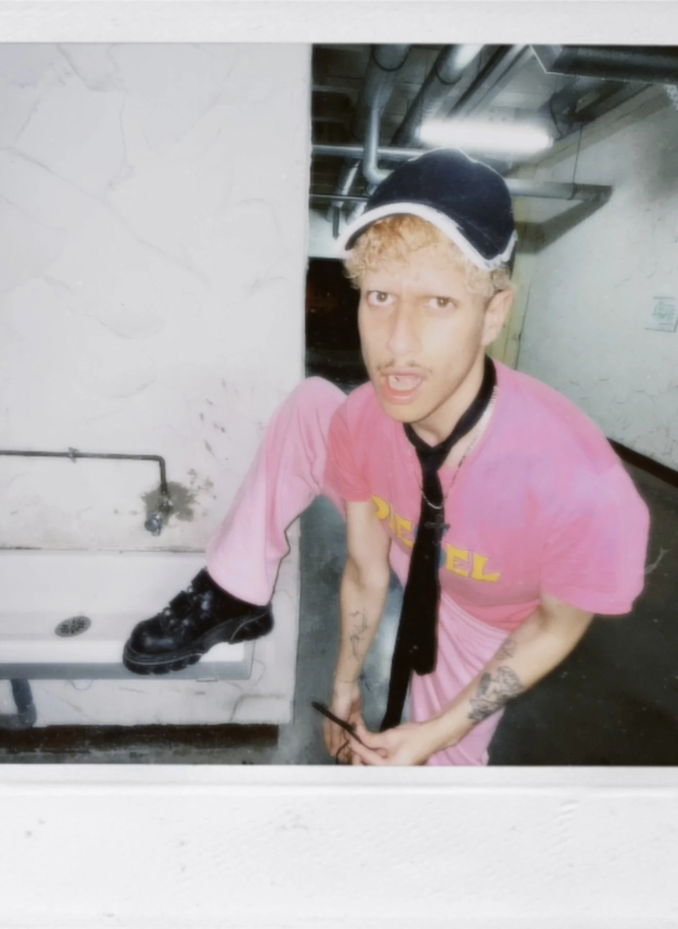 Jeune homme avec des tatouages, portant un chapeau noir et une chemise rose avec une cravate noire, se tenant dans un endroit industriel avec des tuyaux au plafond, prenant un selfie dans un miroir.