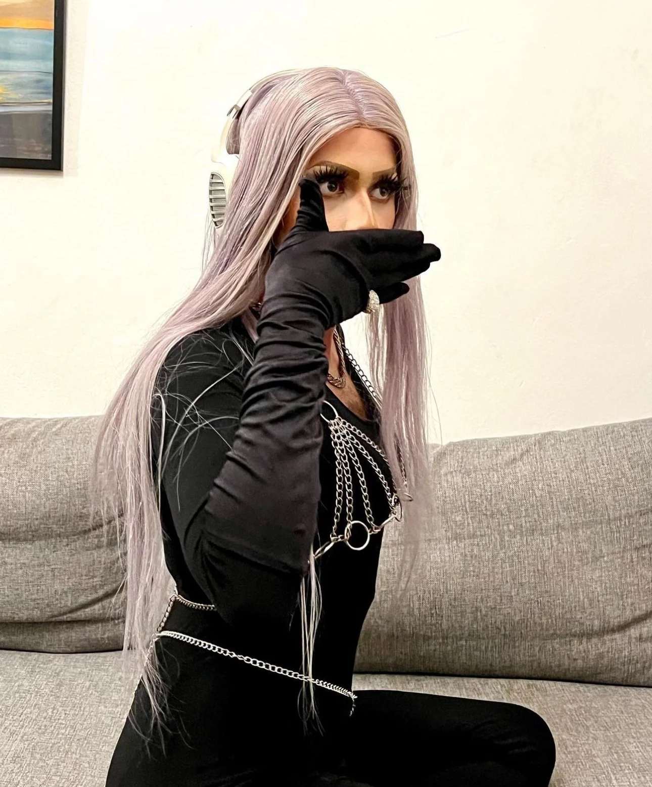 Une femme avec des cheveux longs et argentés portant des vêtements noirs, des gants noirs et des accessoires en chaînes, assise sur un canapé beige, faisant un signe de main près du visage.
