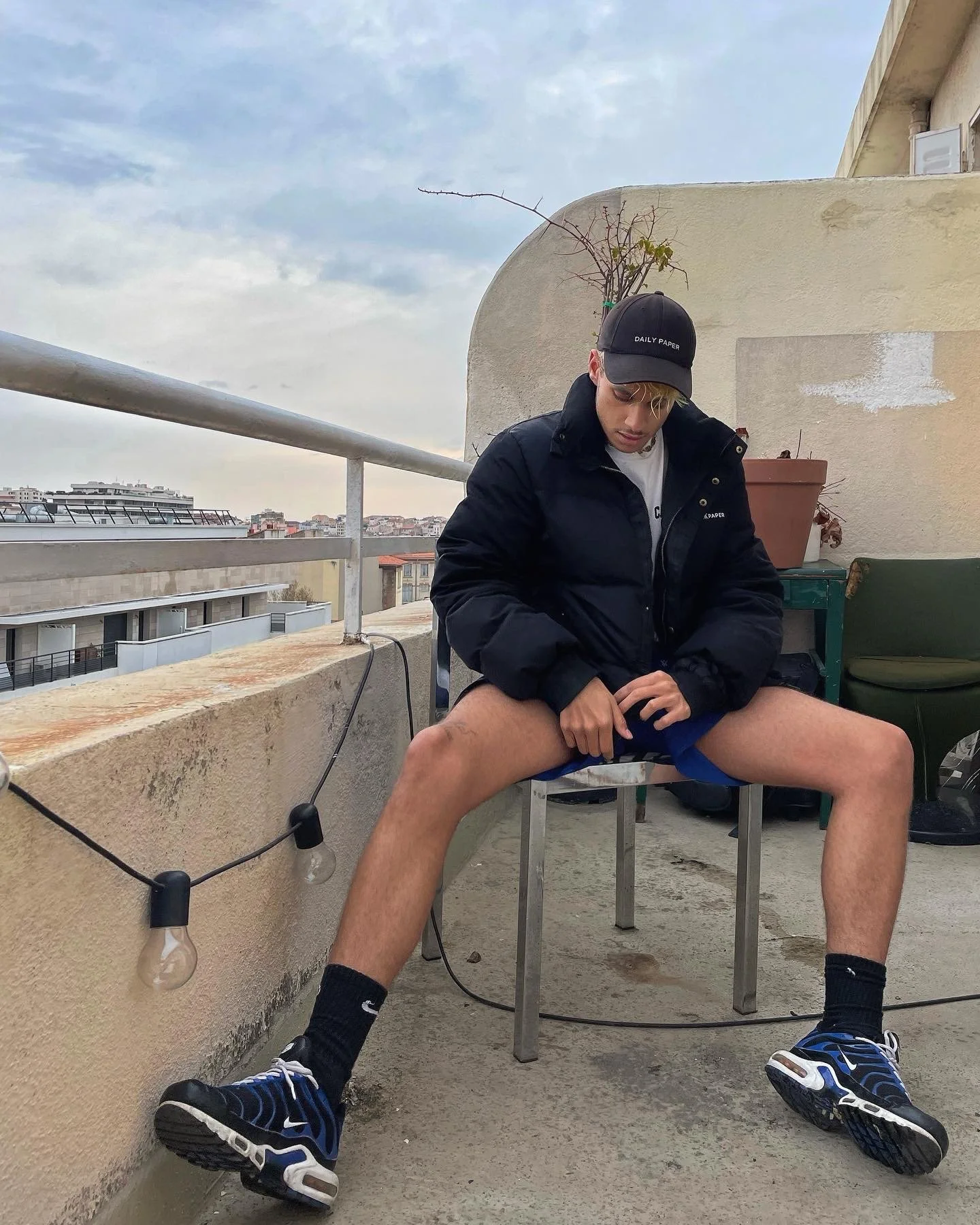 Jeune homme assis sur une chaise, portant un casque noir et un manteau noir, avec les jambes étendues en arrière, sur un balcon avec des décorations de guirlandes lumineuses, un pot de fleurs et une vue urbaine en arrière-plan.