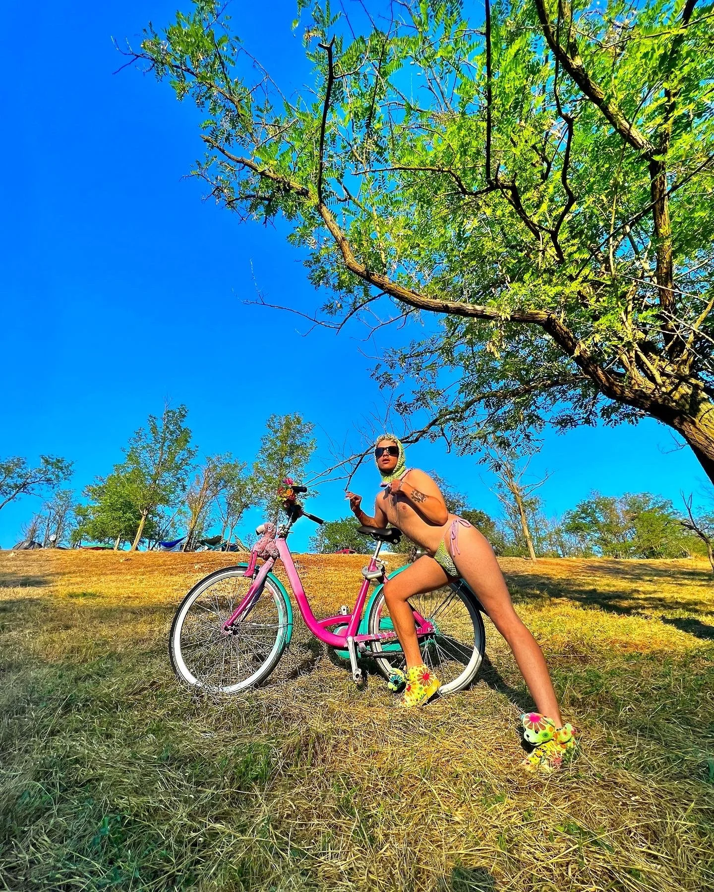 Femme en maillot de bain, lunettes de soleil, avec des chaussures de sport décorées, posant avec un vélo rose dans un parc en plein air sous un ciel bleu avec des arbres verts.