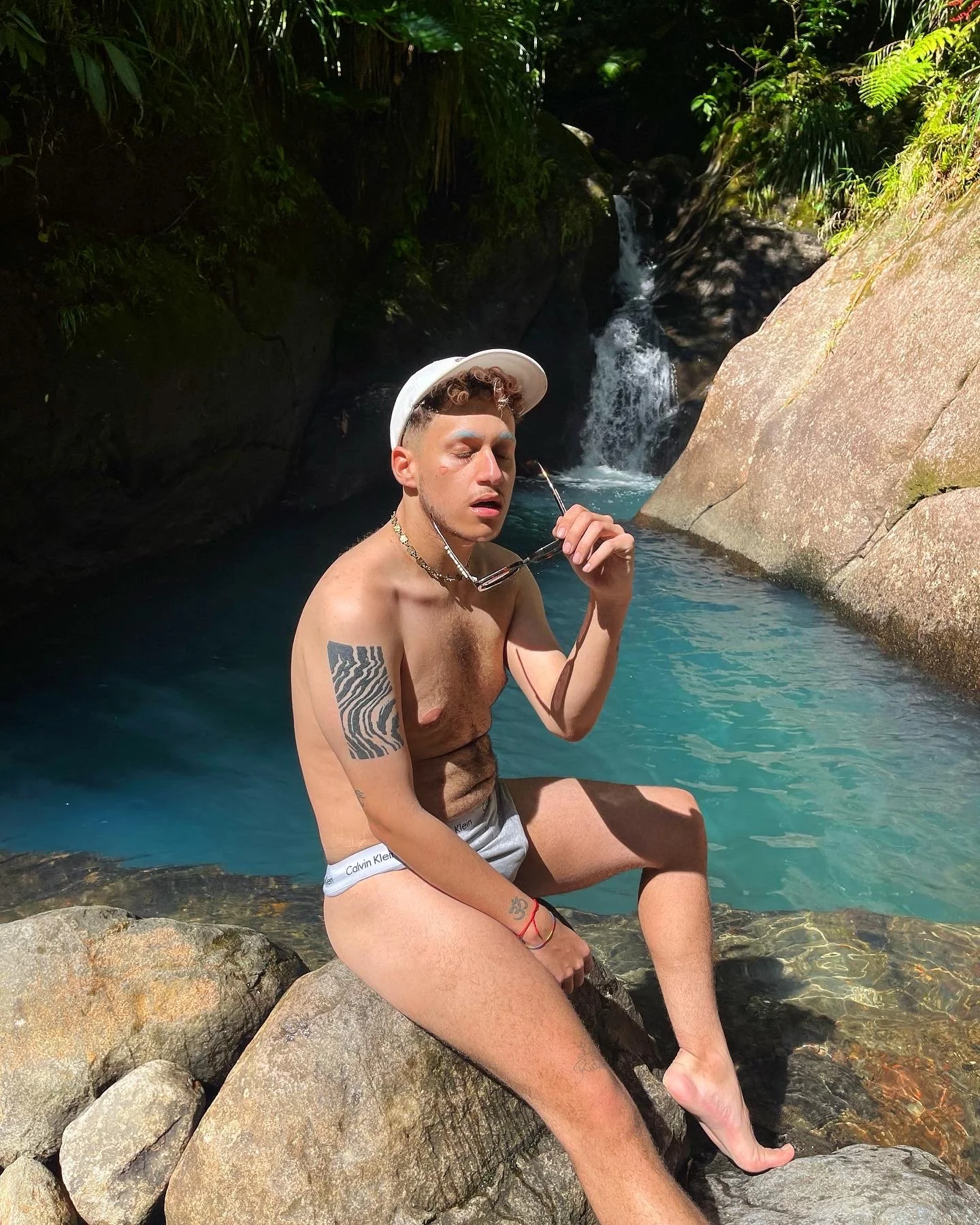 Jeune homme torse nu portant un bonnet blanc, assis sur une roche près d'une cascade dans un environnement naturel, avec des tatouages et des accessoires de mode.