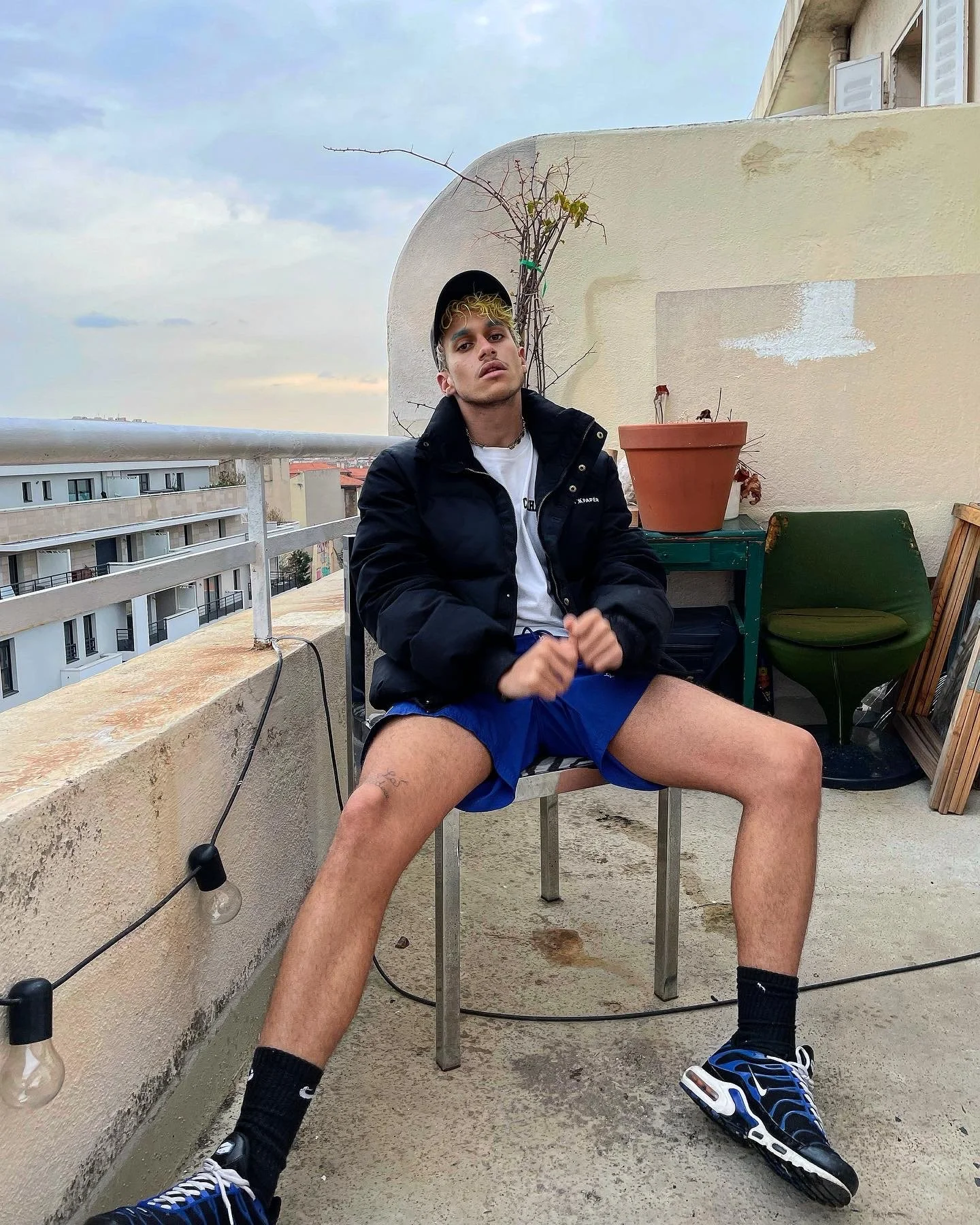 Jeune homme assis sur une terrasse, portant un bonnet, une veste noire et un short bleu, avec des chaussures de sport bleues, un tatouage sur la cuisse gauche, dans un cadre urbain avec un canapé vert et une pot de fleurs derrière lui.