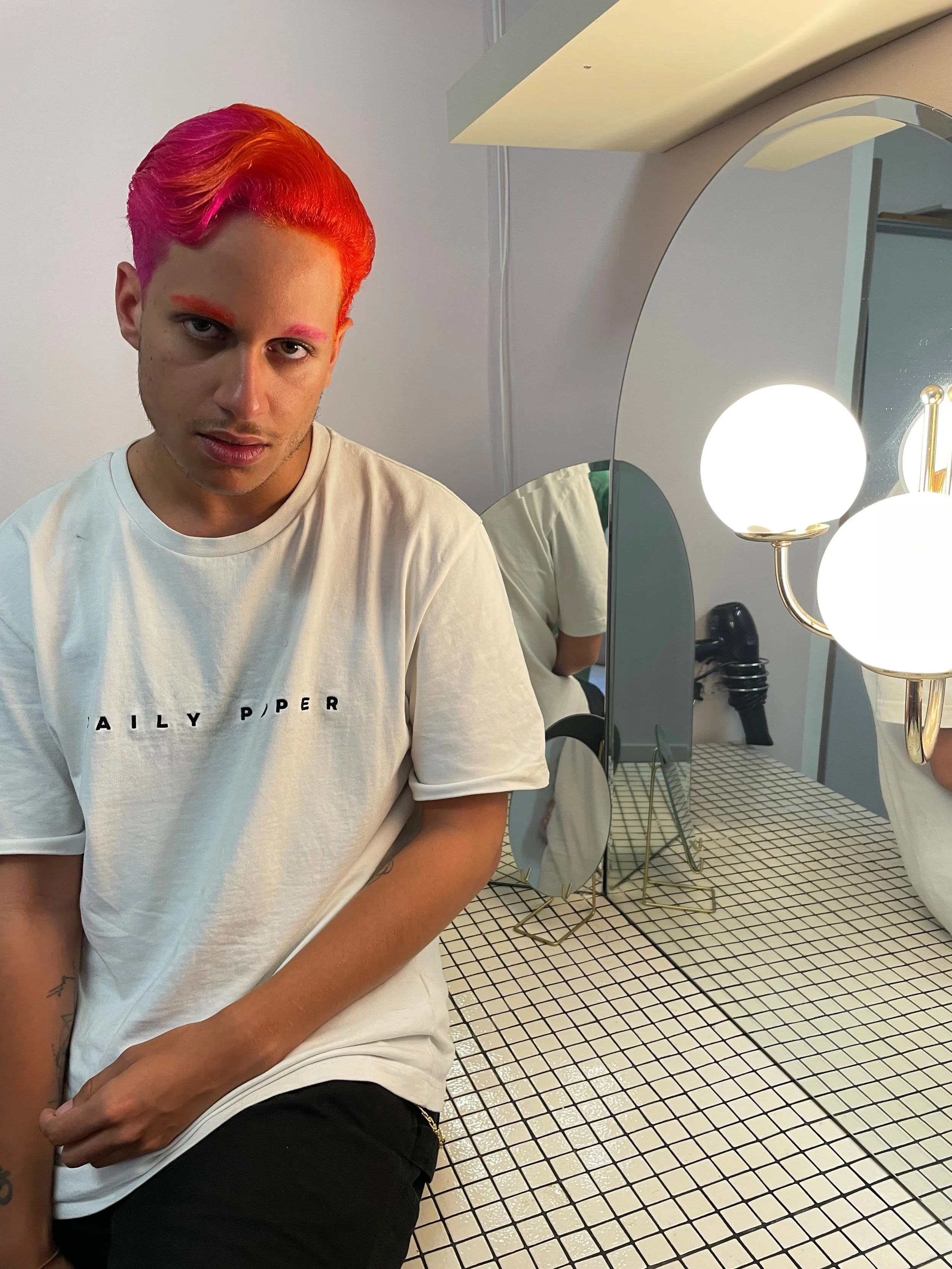 Un jeune homme avec des cheveux roses et rouges assis devant un miroir avec une lampe, portant un t-shirt blanc.
