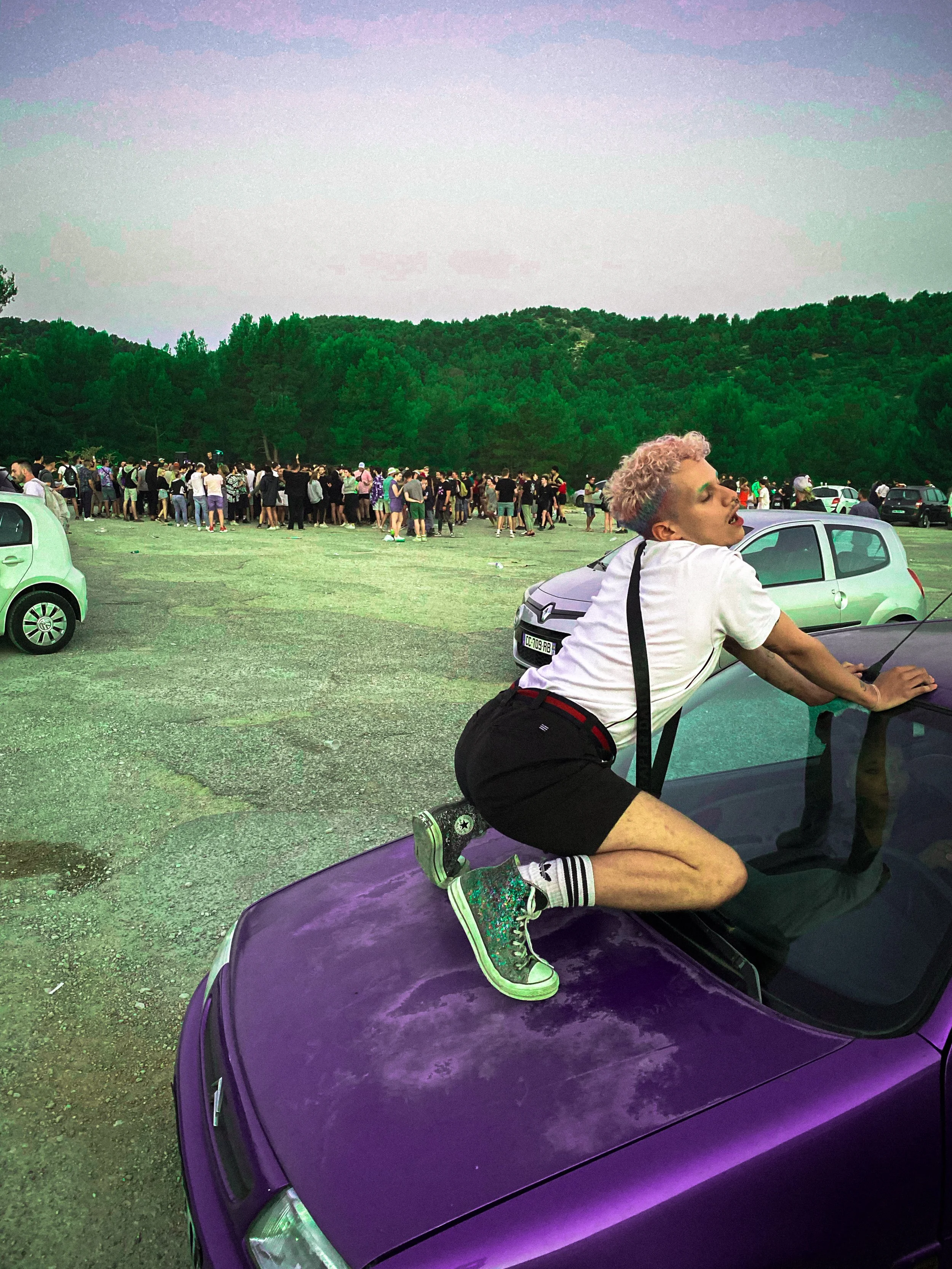 Une personne avec des cheveux bouclés et colorés posant sur une voiture violette dans un parking en regardant vers la droite, avec un groupe de personnes au fond et une forêt derrière.