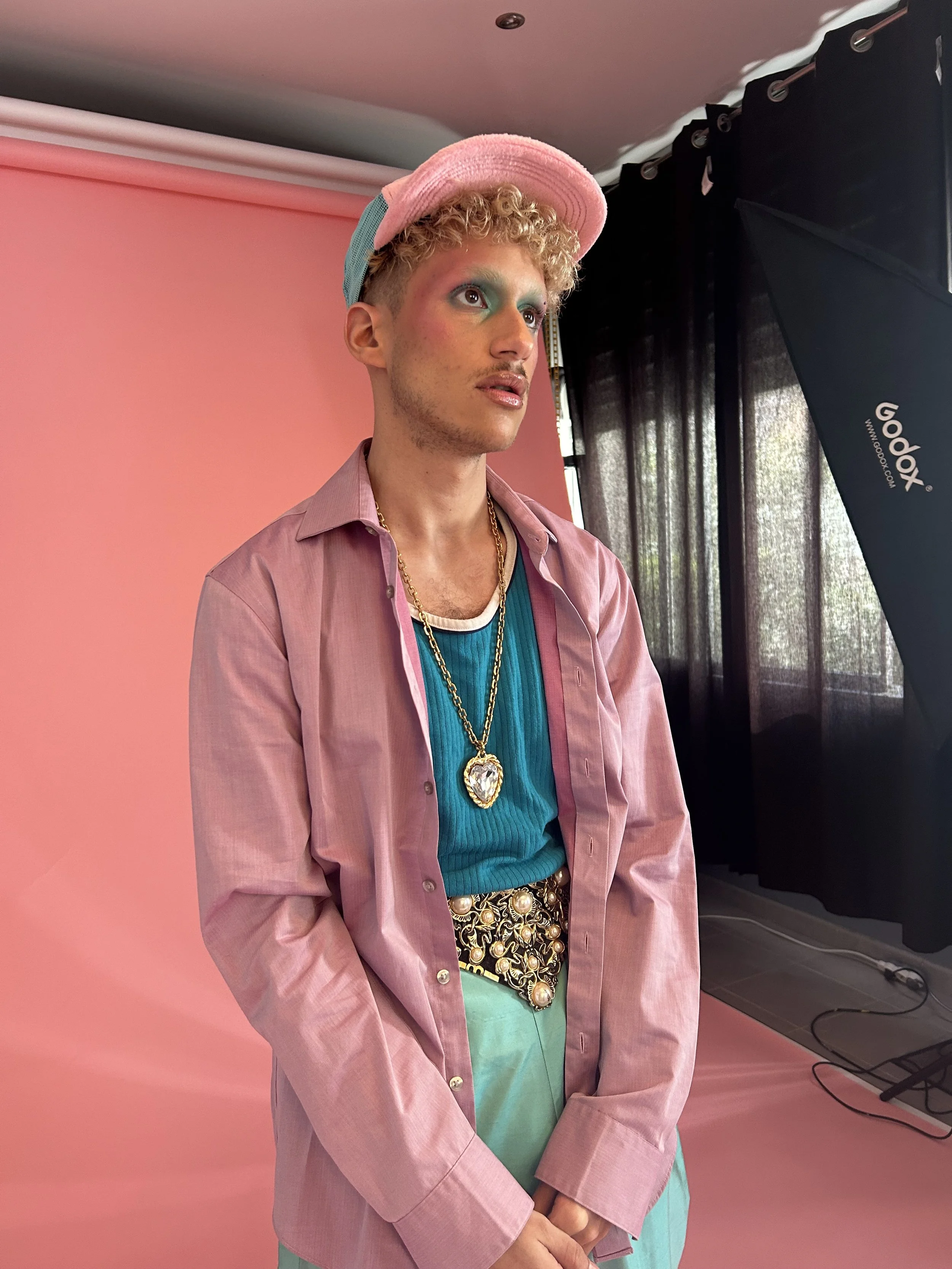 Jeune homme avec maquillage coloré, portant un bonnet rose, une chemise rose sur un t-shirt turquoise, une ceinture ornée de bijoux, photoshoot dans un studio avec fond rose et panneau lumineux.