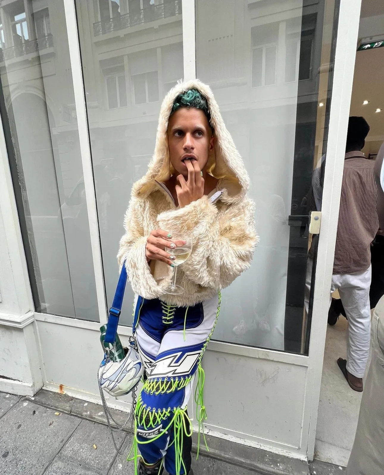Jeune homme avec une veste en peluche beige, pantalons à motifs colorés, tenant une coupe de vin, à l'extérieur d'un bâtiment.