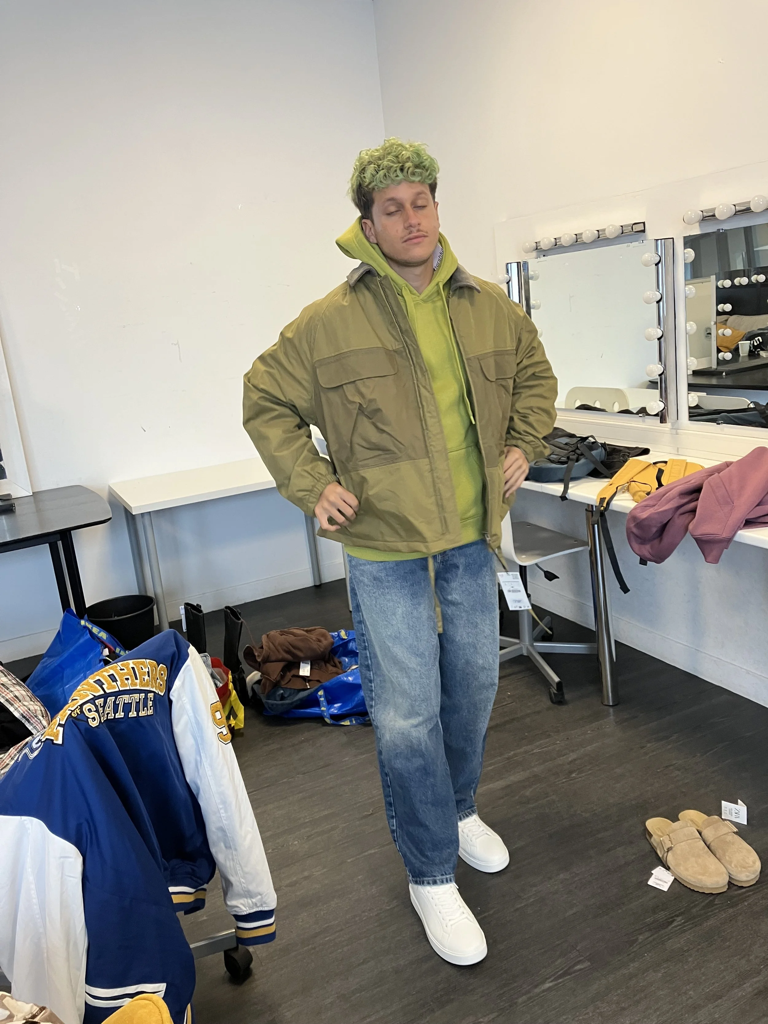 Jeune homme avec des cheveux bouclés verts, portant un sweat vert fluo sous une veste khaki, jeans et chaussures blanches, dans une pièce avec des miroirs et des vêtements
