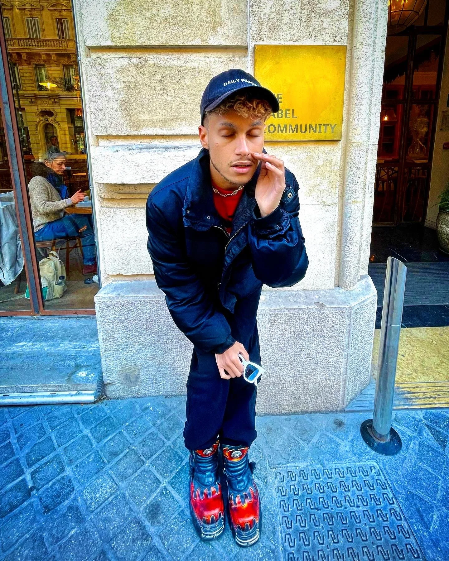 Un jeune homme debout devant un mur en pierre, portant une casquette noire, une veste noire, des bottes à motif de flammes rouges et bleues, tenant des lunettes de soleil blanches et rouges, avec un doigt sur le visage, à côté d'un panneau jaune avec