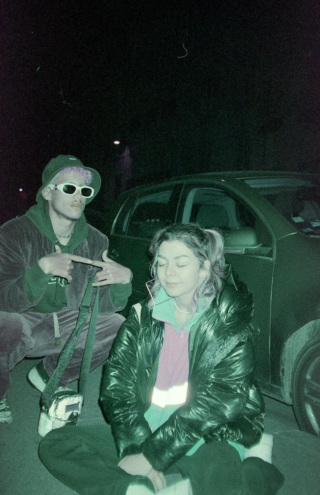 Deux jeunes adultes posent avec un véhicule en arrière-plan dans un environnement sombre. La personne à gauche porte des lunettes de soleil blanches, un chapeau, et un sweat à capuche vert. La personne à droite, assise par terre, a des cheveux en que