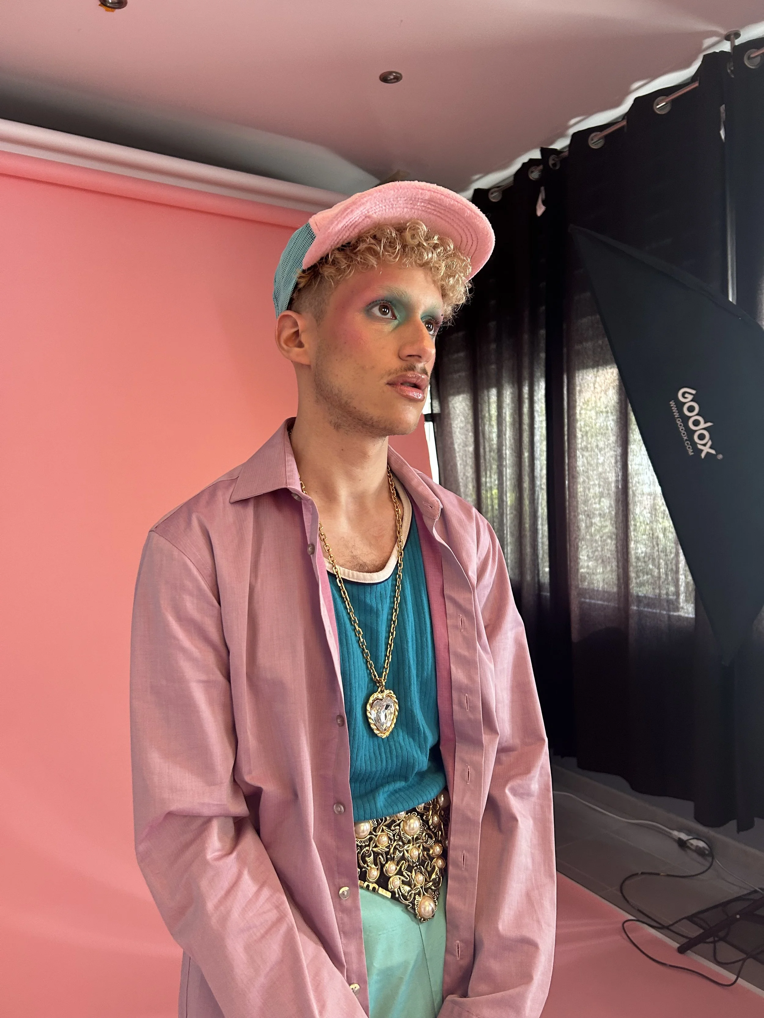 Person posant dans un studio photo, portant un chapeau pastel, une veste rose, un t-shirt turquoise, et des bijoux, avec un fond rose et un éclairage professionnel.