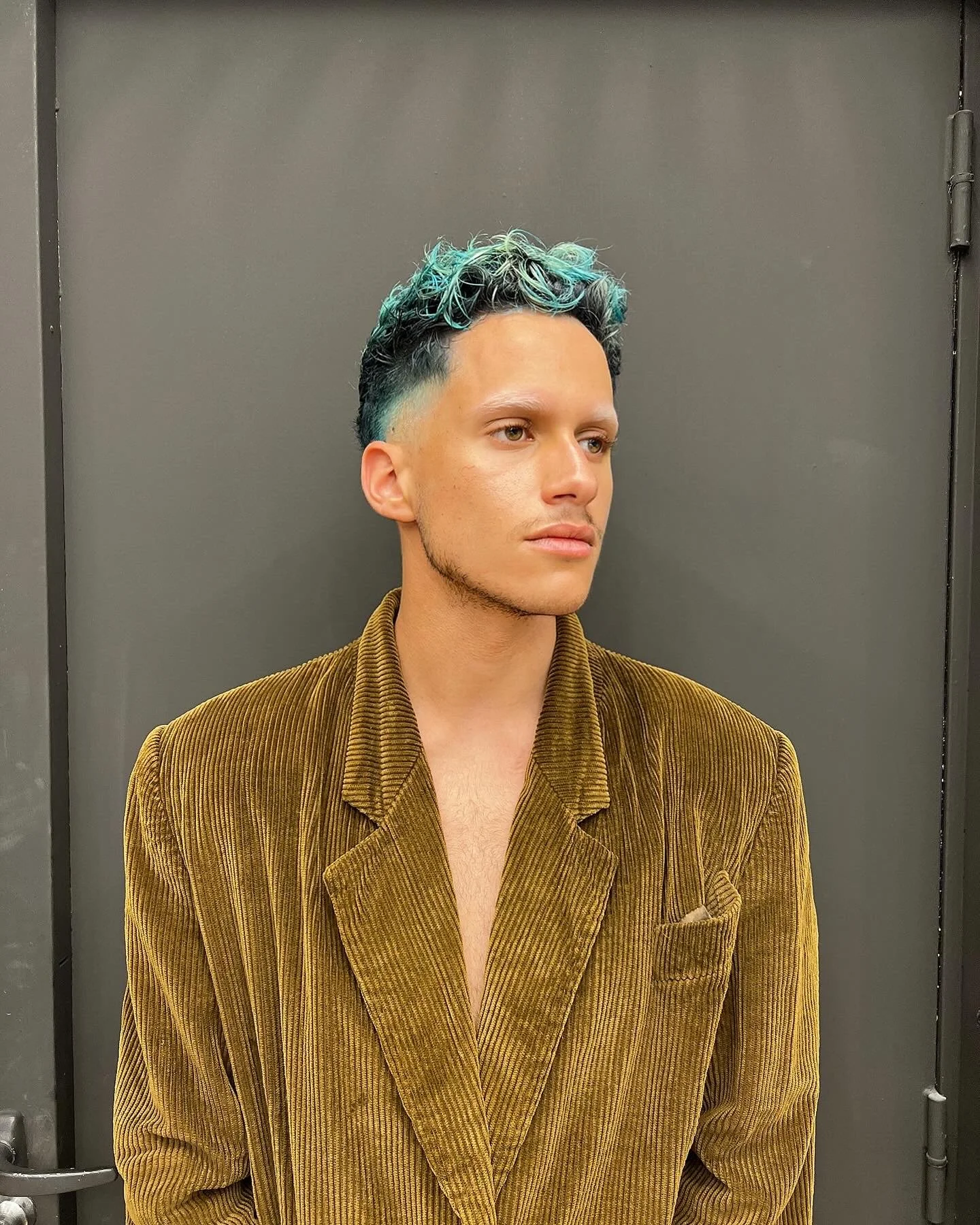 Un jeune homme avec des cheveux en partie teints en bleu, portant une veste en velours marron clair, debout contre un mur gris.