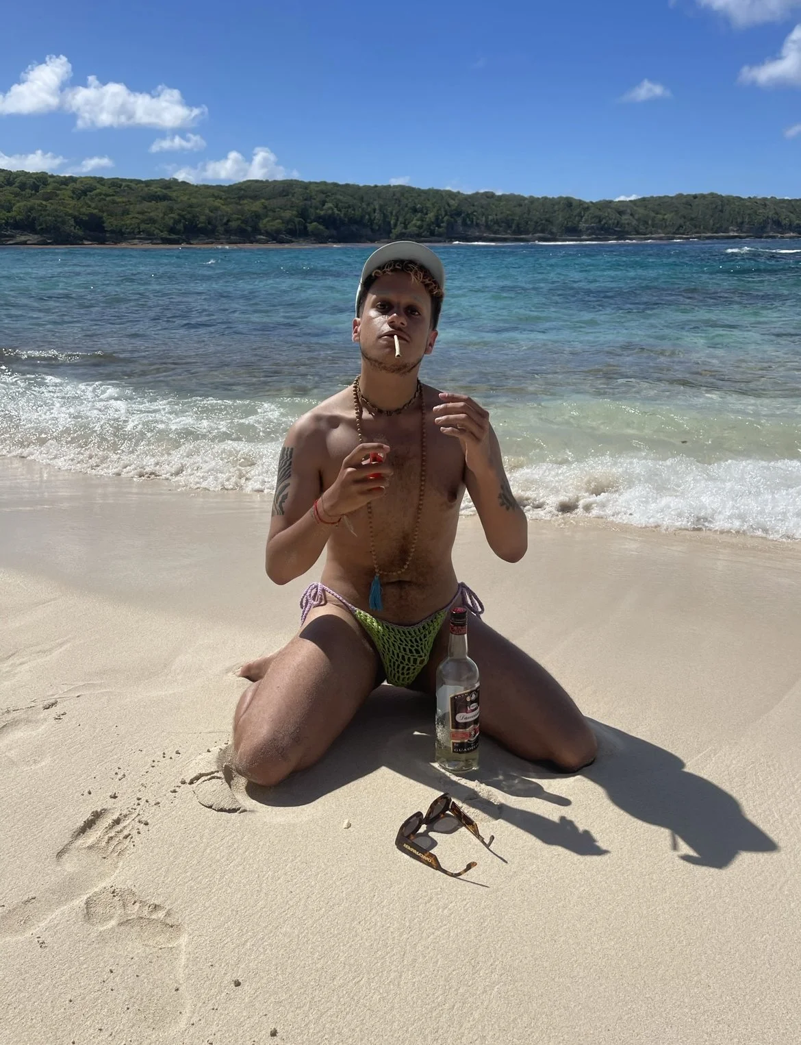 Jeune homme assis à genoux sur la plage, portant un chapeau blanc, une chaîne et un bracelet, avec une bouteille d'alcool, des lunettes de soleil, et un cigare dans la bouche, plage rocheuse et mer en arrière-plan.