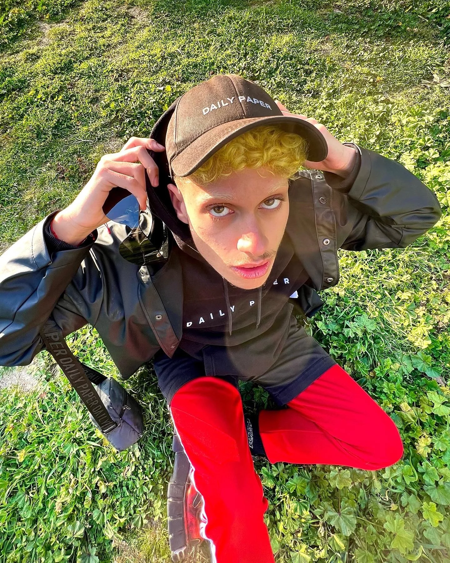 Jeune homme avec des cheveux blonds bouclés portant une casquette marron, une veste noire, un T-shirt noir et des pantalons rouges, prenant un selfie en plein air sur un fond de végétation verte.