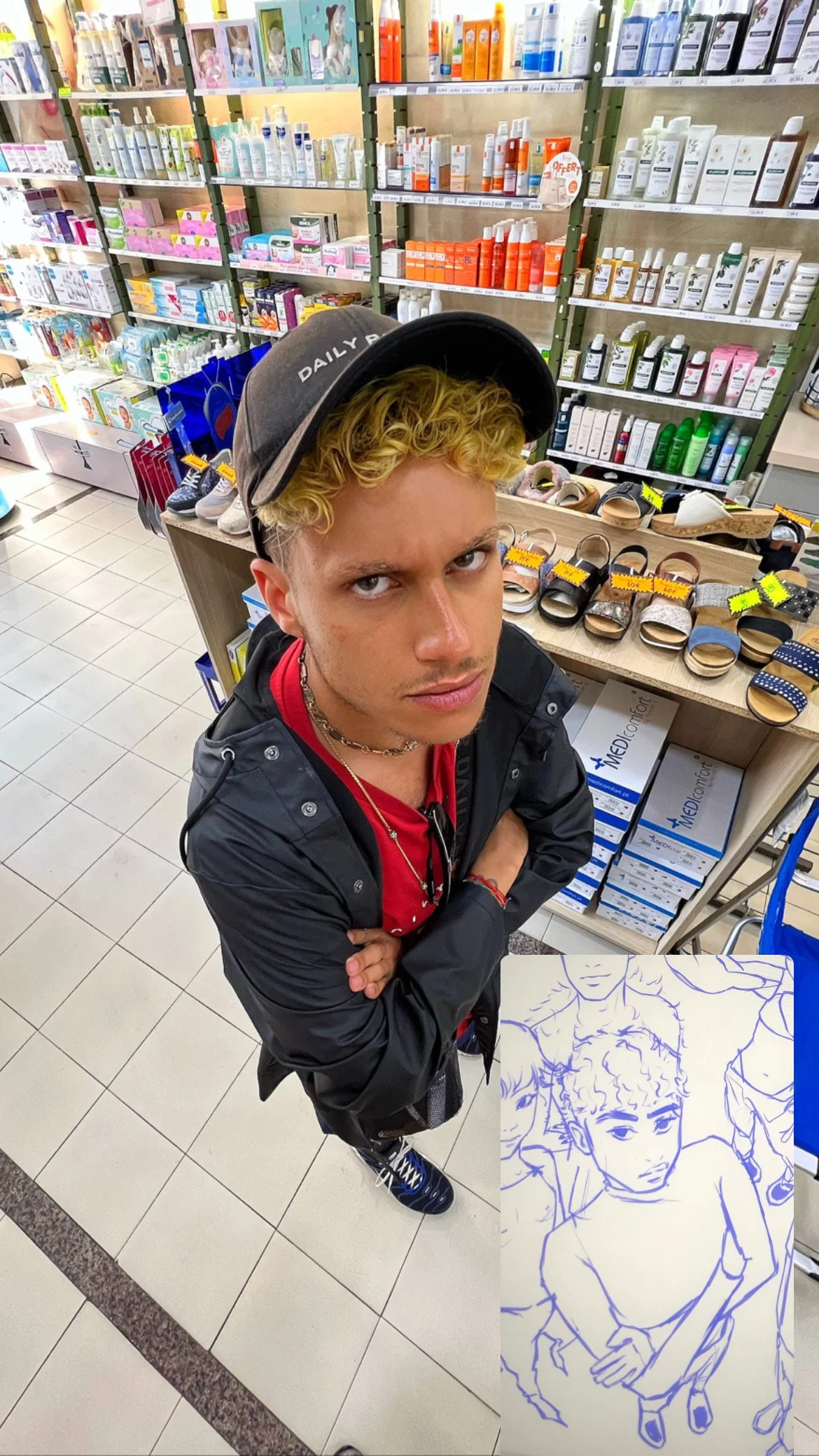 Un jeune homme avec des cheveux bouclés, portant une casquette et une veste noire, se tient dans une pharmacie ou un magasin de produits cosmétiques. Il regarde fixement l'appareil photo, bras croisés, avec en arrière-plan des étagères remplies de pr