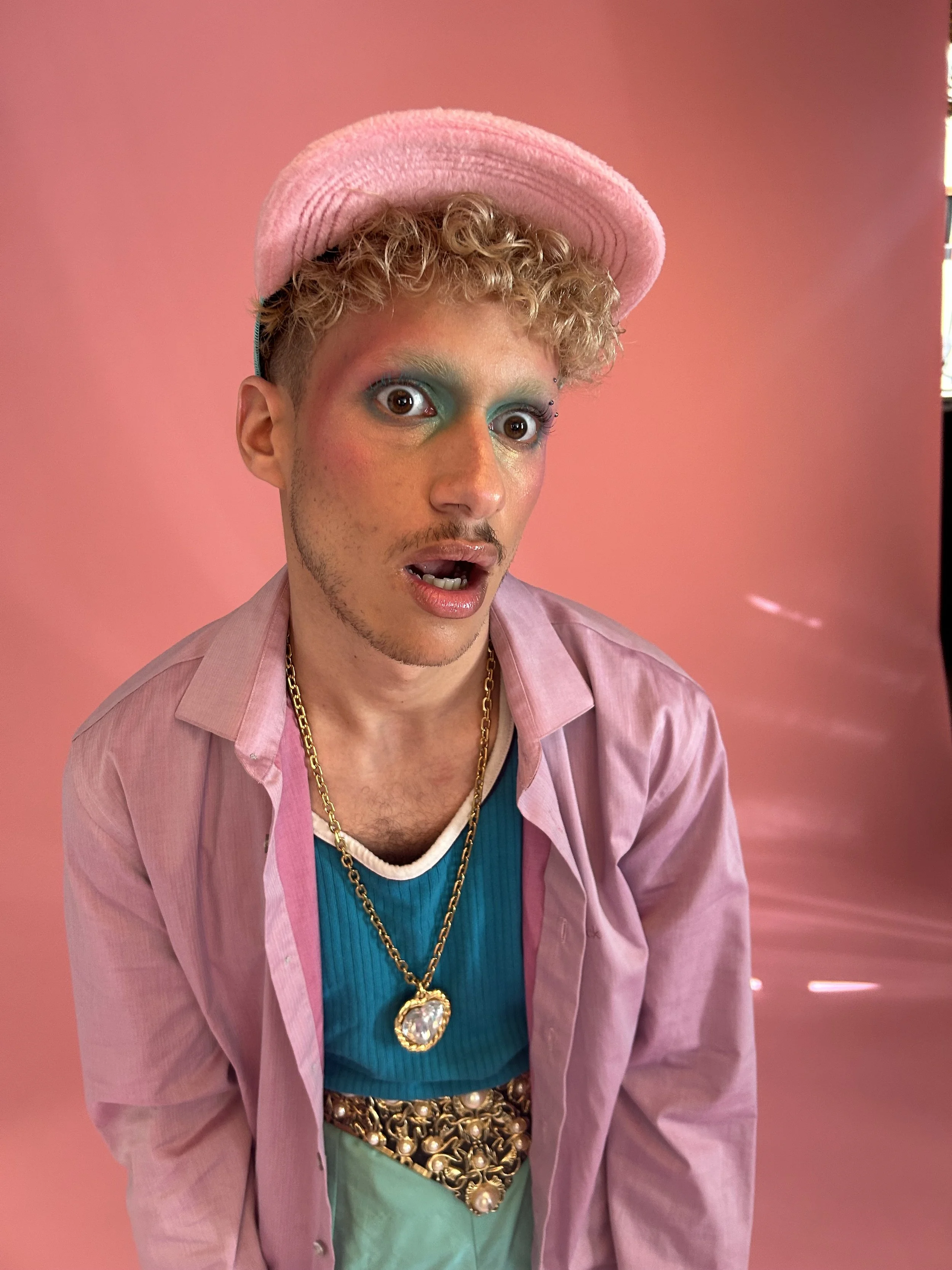 Une personne portant un chapeau rose, un maquillage coloré avec des ombres à paupières bleu et vert, un haut en cachemire bleu, une veste rose, une chaîne en or avec un pendentif en forme de cœur, et une ceinture dorée avec des perles, posant devant