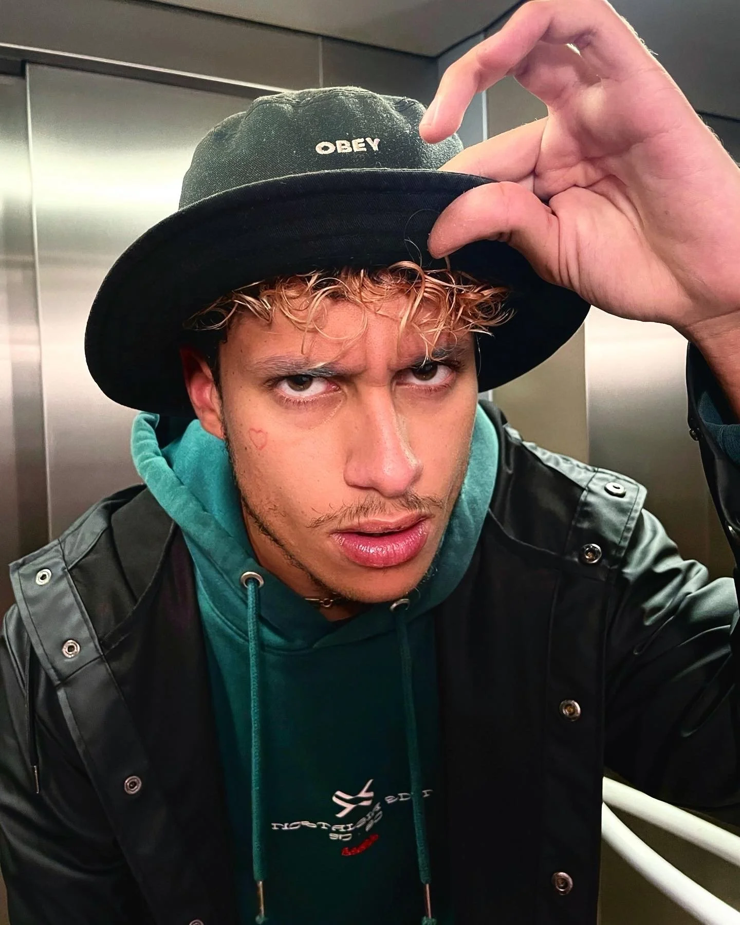 Jeune homme avec des cheveux bouclés blonds, portant un chapeau noir avec ''OBEY'' écrit dessus, un hoodie vert et une veste en cuir, regardant intensément.