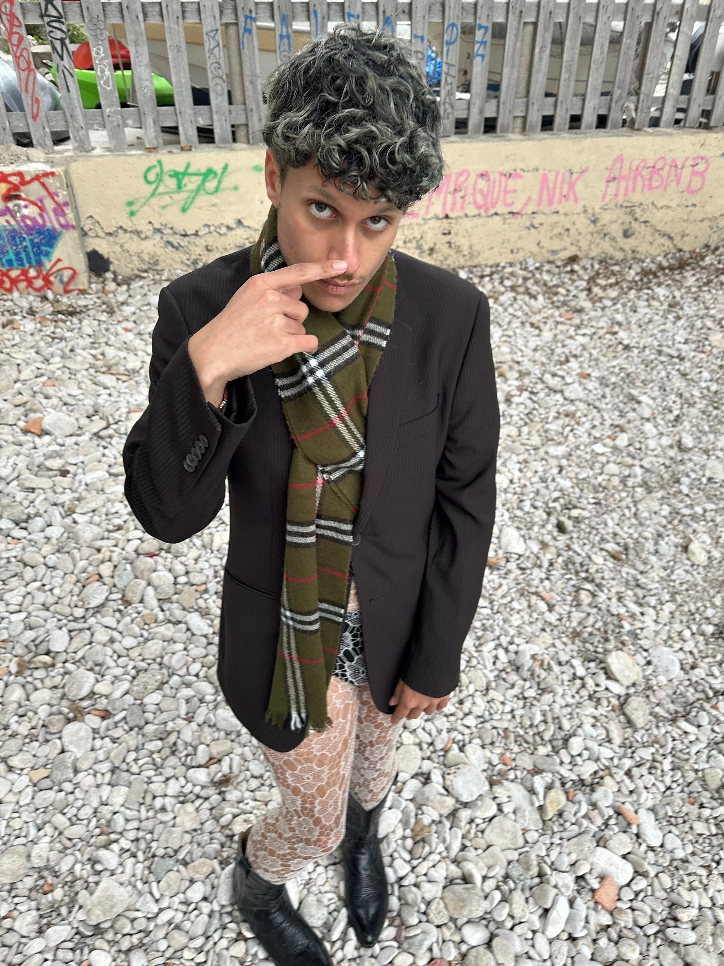 Jeune homme en costume noir, foulard à carreaux, chaussettes en dentelle, bottines noires, posant sur un fond de mur avec graffiti, piétinant un sol de pierres.