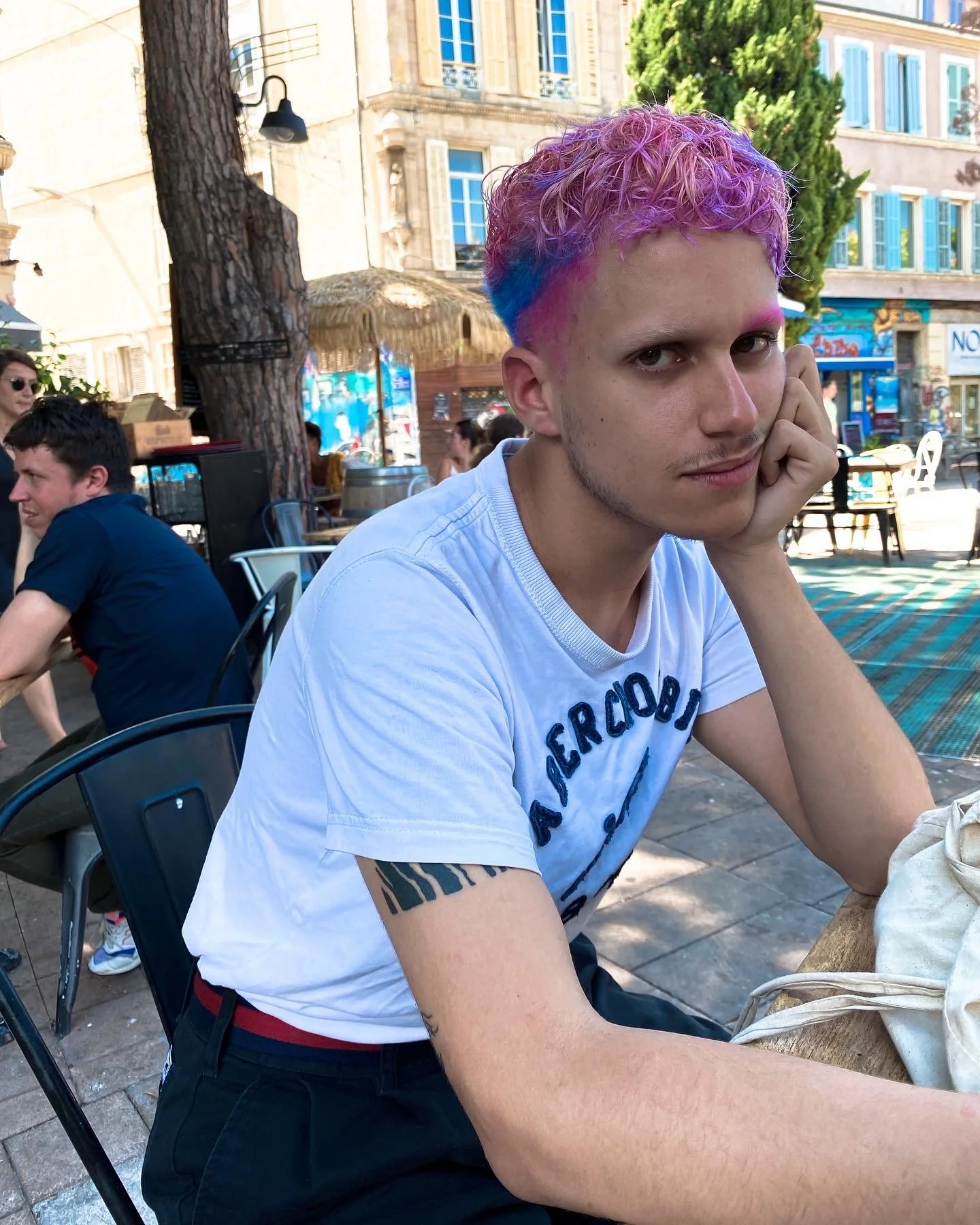 Un jeune homme aux cheveux bouclés et colorés en rose, violet et bleu, assis à une table en plein air dans une ville. Il porte un t-shirt blanc avec le logo Aeropostale, regarde fixement l'appareil photo et repose sa joue sur sa main. En arrière-plan