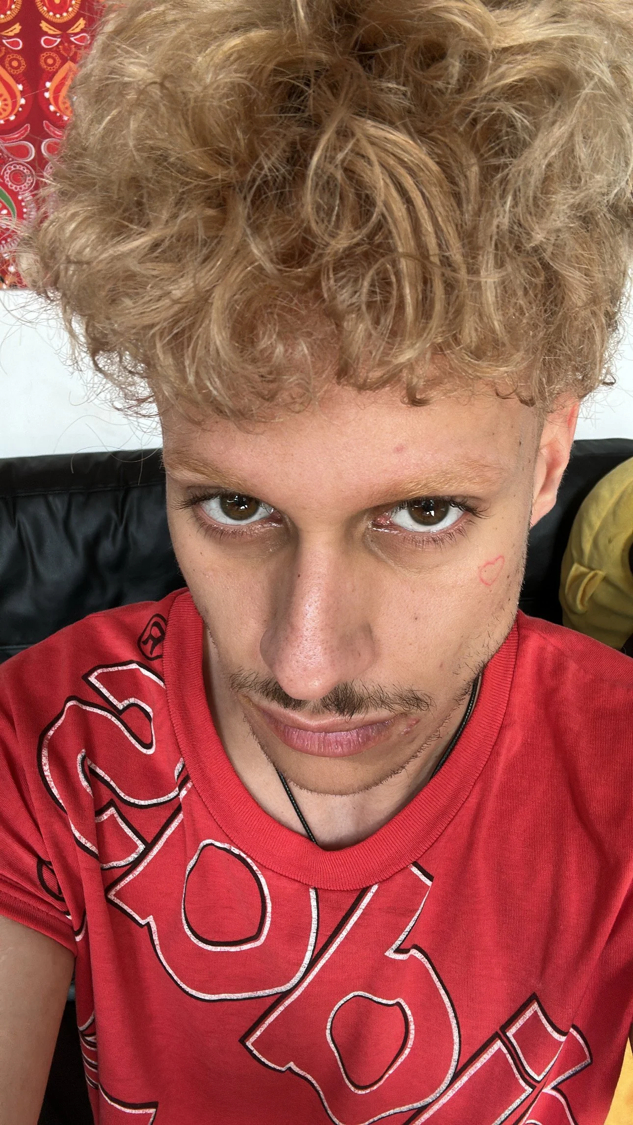 Jeune homme aux cheveux bouclés et blondissants, portant un t-shirt rouge avec un gros motif blanc, assis sur un canapé noir.