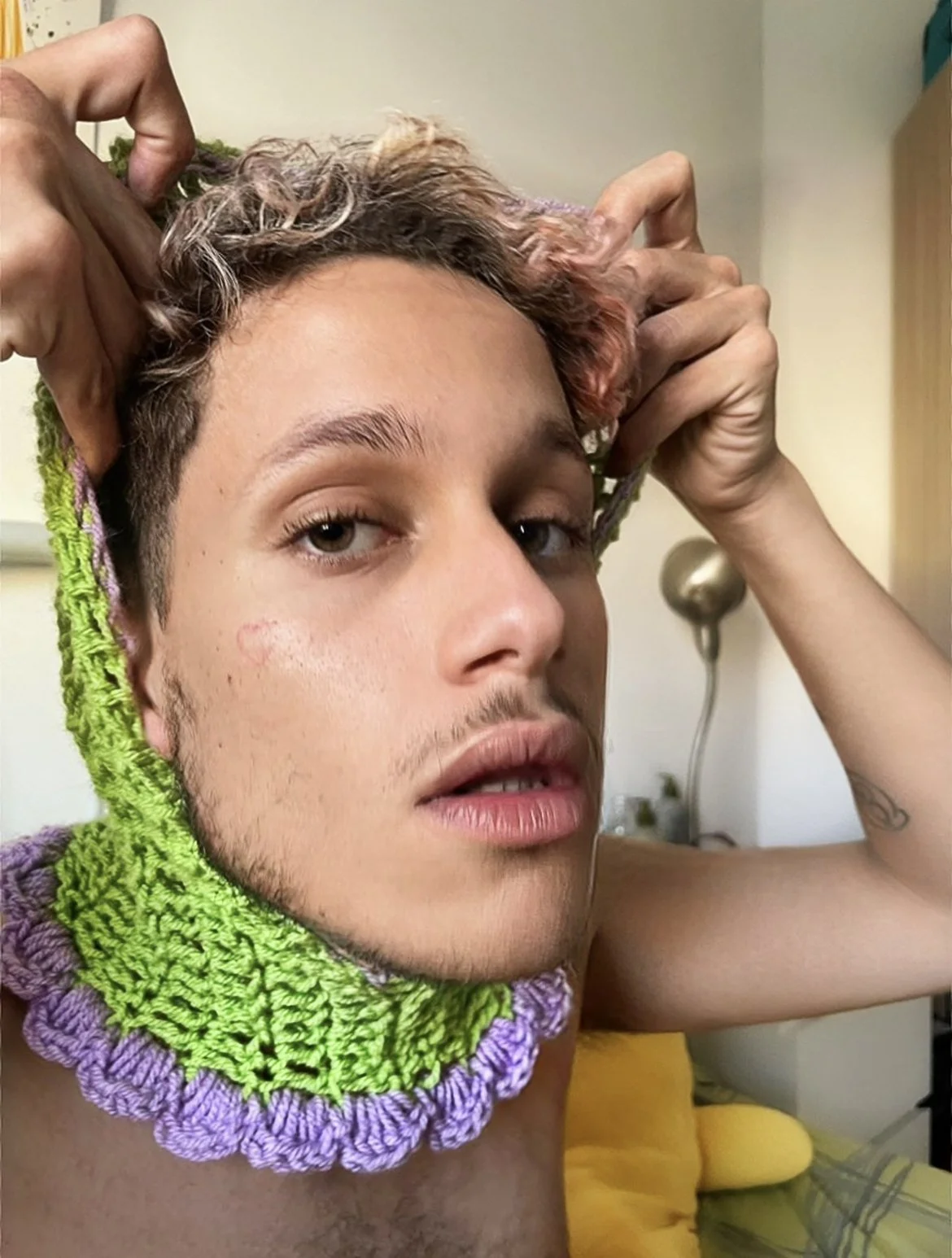 Jeune homme portant un collier en crochet vert et violet, posant avec ses mains derrière la tête, dans une pièce devant un lampadaire