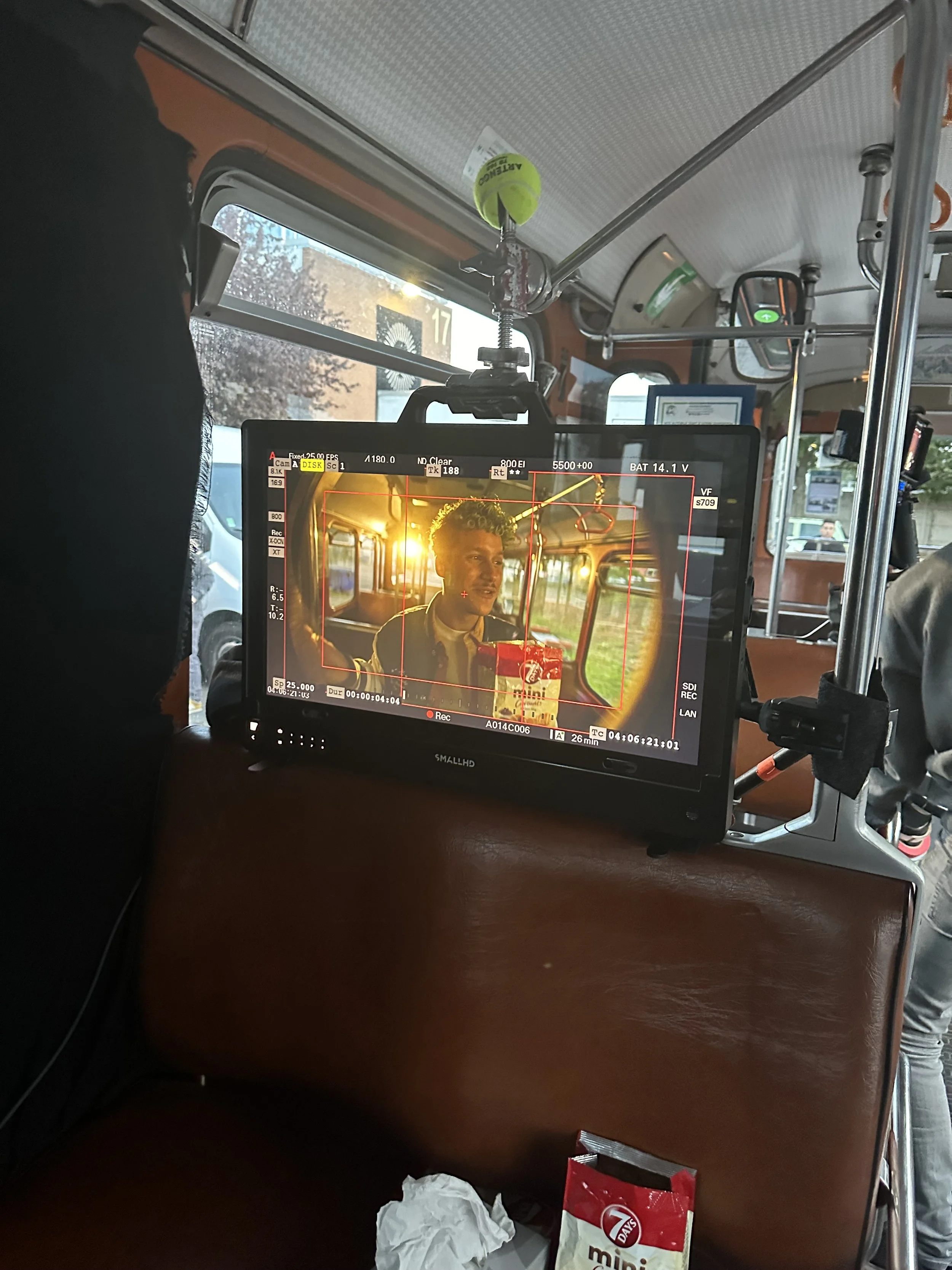 Écran de caméra de tournage montrant un jeune homme avec des cheveux bouclés dans un bus durant un coucher de soleil, portant une veste et une chemise, avec un snack sur ses genoux.