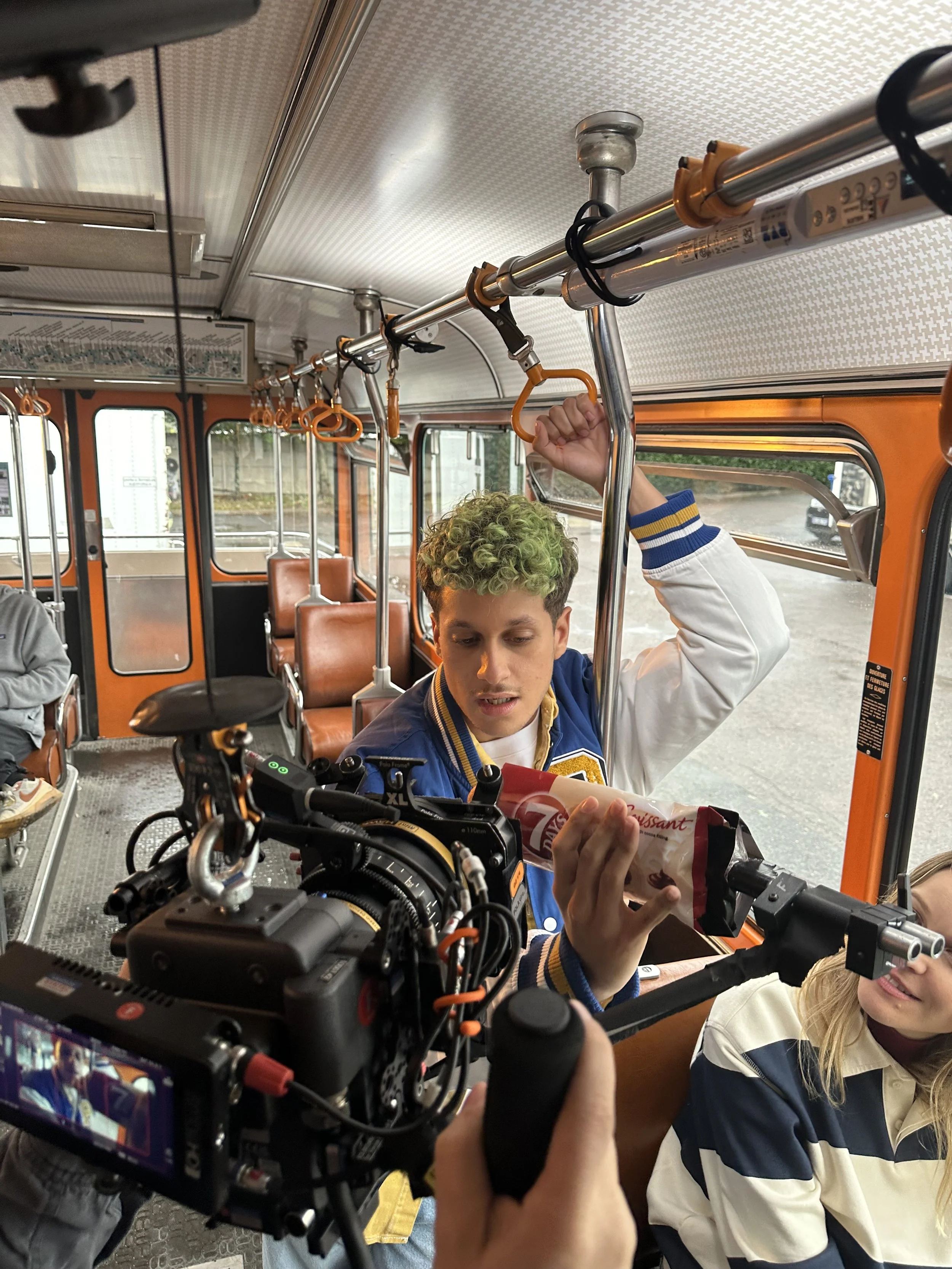 Jeune homme avec des cheveux bouclés verts, portant un blouson bleu et blanc, tenant un paquet de biscuits dans un bus orange antique, avec une caméra en premier plan et une femme à ses côtés.