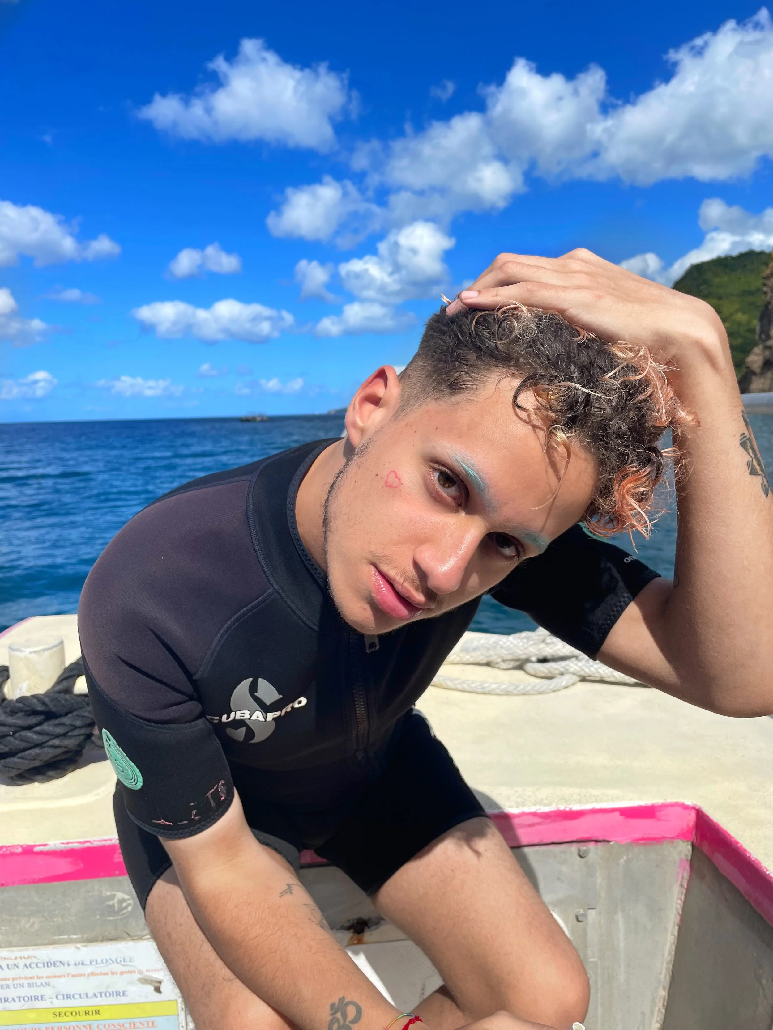 Jeune homme inclinée sur un bateau en mer, souriant, avec ciel bleu, nuages, et côte en arrière-plan.