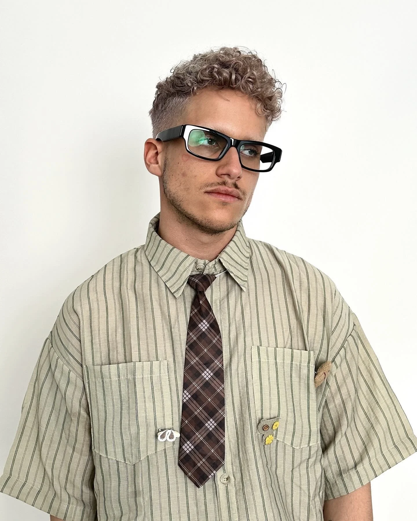 Jeune homme portant des lunettes épaisses, une chemise à rayures avec des badges et épinglettes, et une cravate en tissu à carreaux, posant devant un mur blanc.