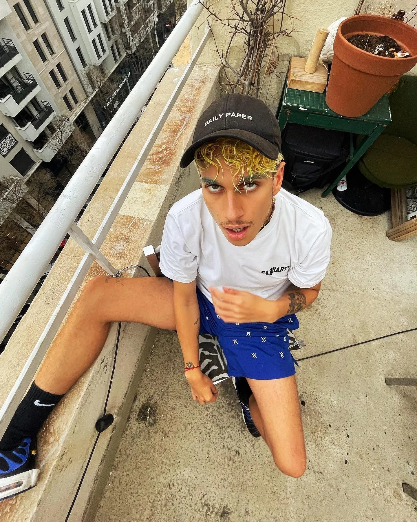 Jeune homme assis sur un balcon, portant une casquette noire avec l'inscription "DAILY PAPER", une chemise blanche, un short bleu, et des chaussures de sport noires et bleues. Il a des cheveux blonds tressés et une expression faciale interrogative. À
