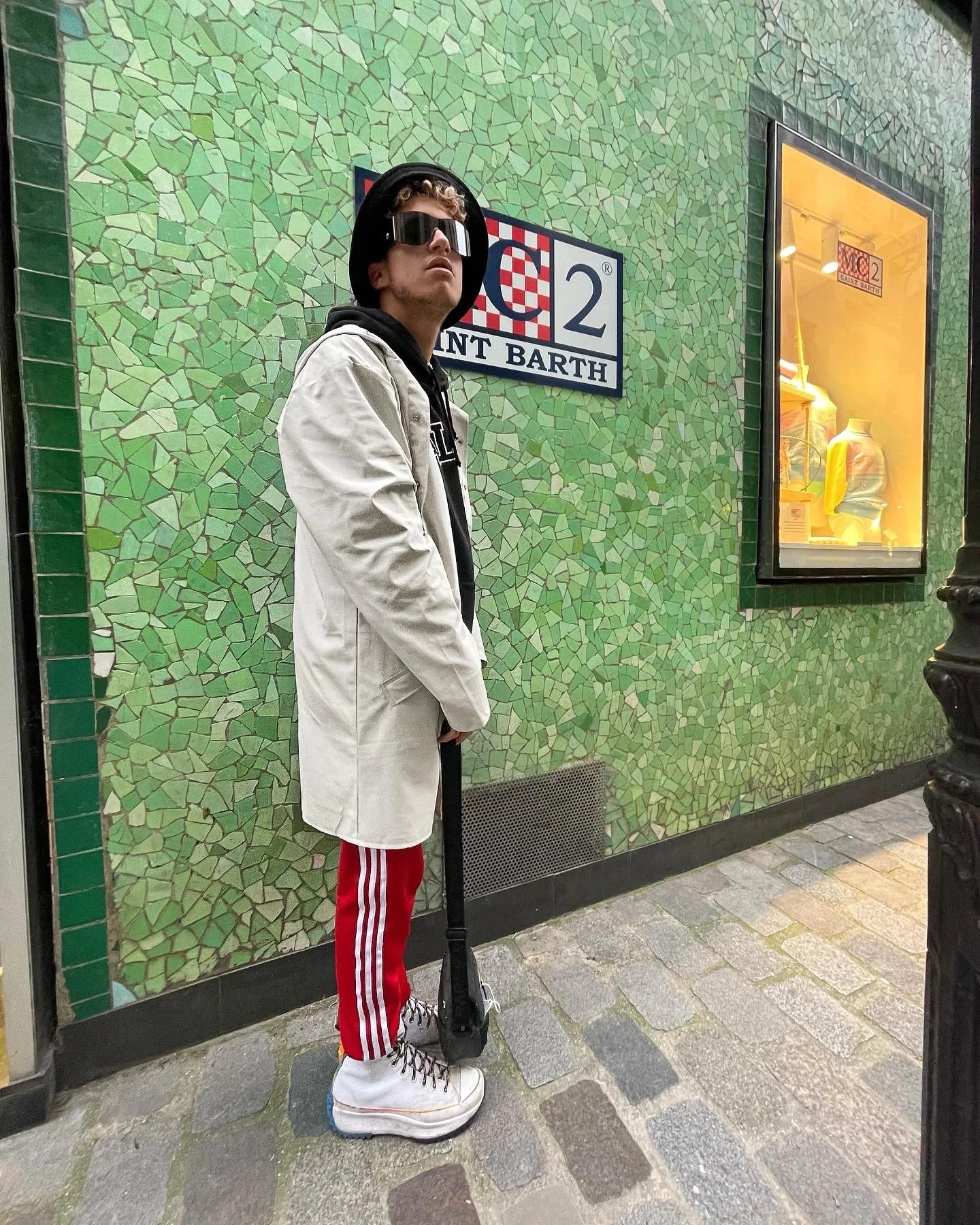 Jeune homme portant un manteau blanc, un hoodie noir, un chapeau noir, des lunettes de soleil, des pantalons rouges à rayures blanches, et des baskets blanches, debout devant un mur en mosaïque verte à Saint-Barthélemy, avec une planche de skateboard