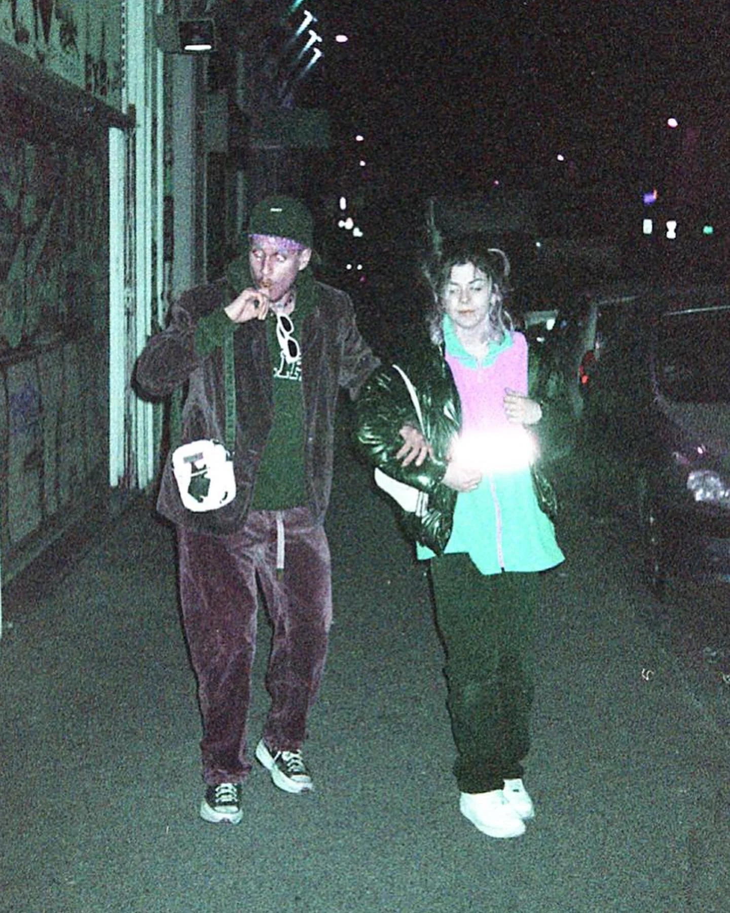 Deux jeunes personnes marchant dans une rue la nuit, l'une portant une veste en cuir noire, et l'autre un sweat coloré, avec des voitures en arrière-plan.