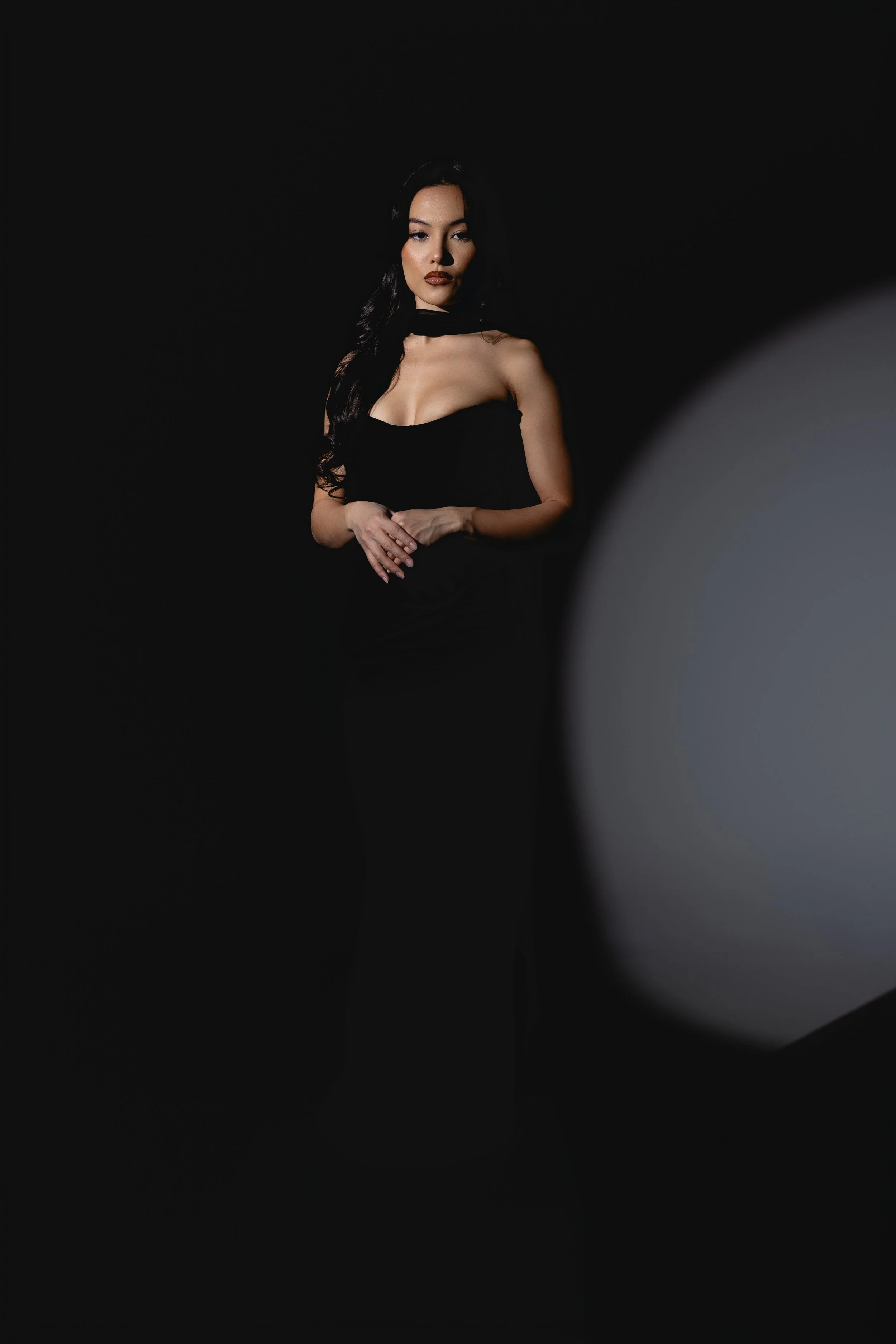A woman with long dark hair, beige skin, and bold makeup, wearing a black sleeveless top and choker, standing against a black background with soft lighting on her face.