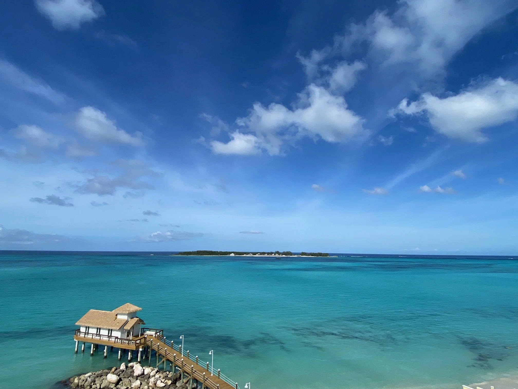 Overwater bungalow with a thatched roof and a wooden walkway extending over turquoise waters toward a small island in the distance under a partly cloudy blue sky.