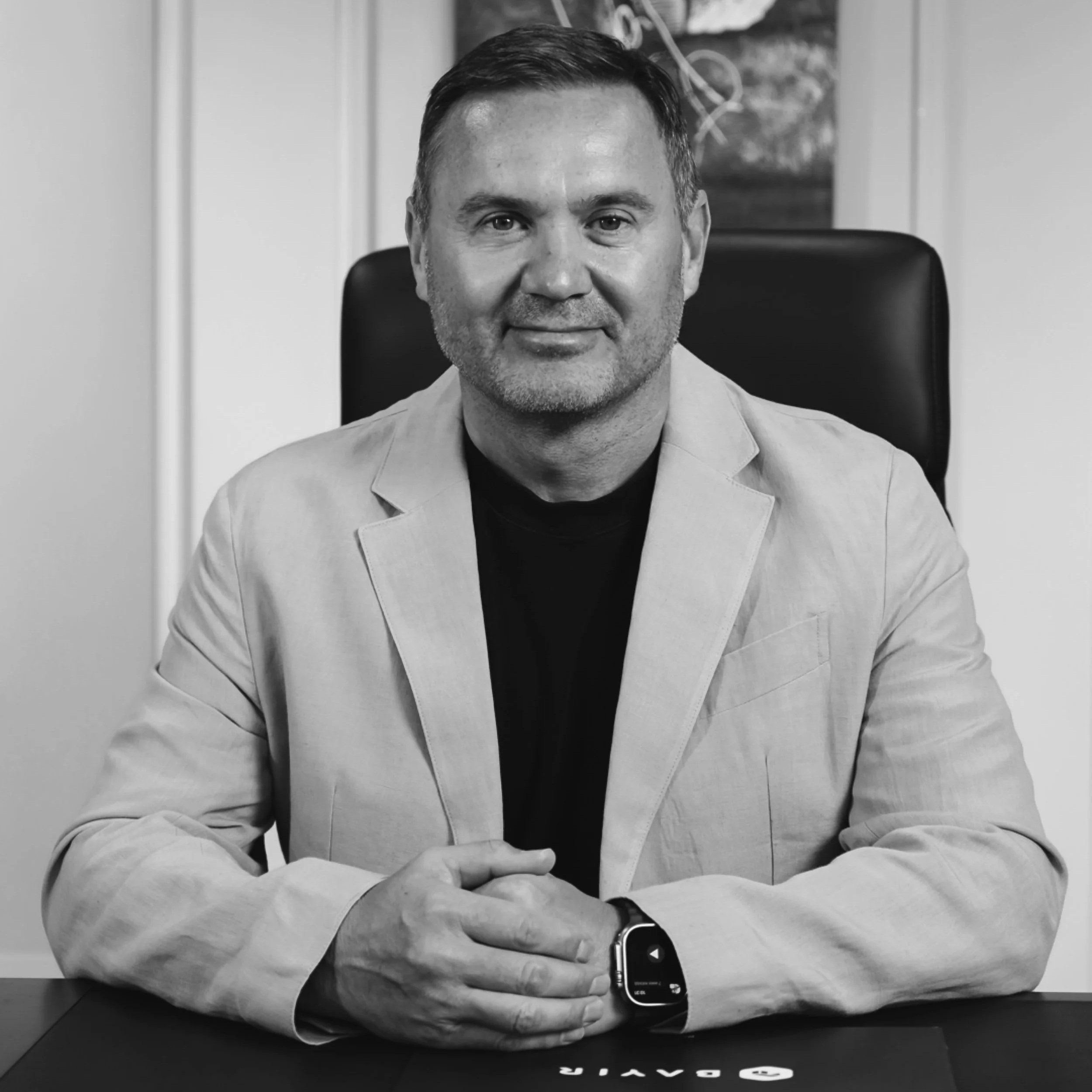 A man sitting at a desk in an office, wearing a light-colored blazer and a smartwatch on his left wrist, with a relaxed and confident expression.