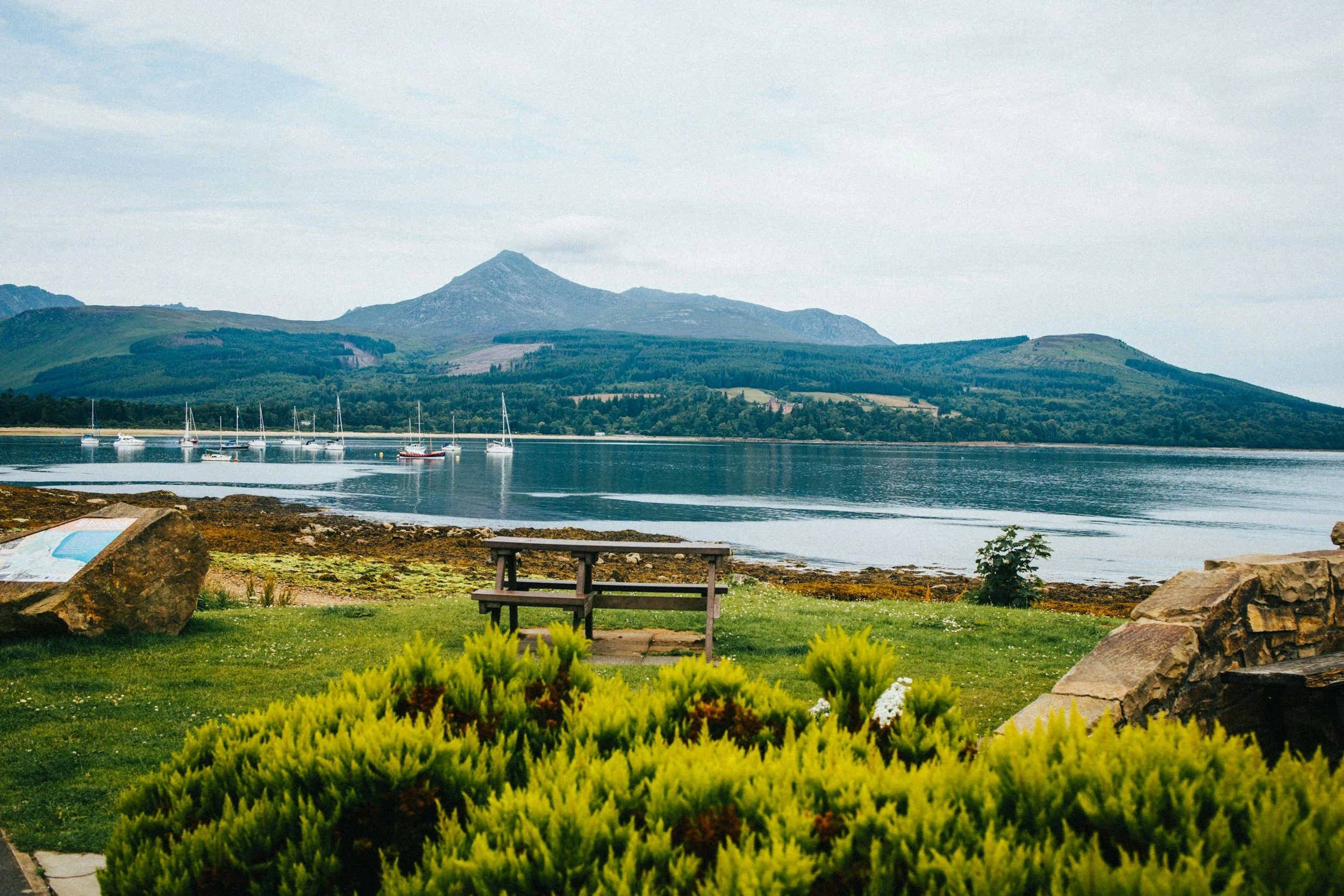 View of an Arran mountain landscape with sea and sailboats in the foreground, green shrubbery, and a cloudy sky.