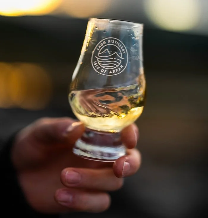 A hand holding a glass of white wine with a logo that says Lago Distillery Isle of Arran.