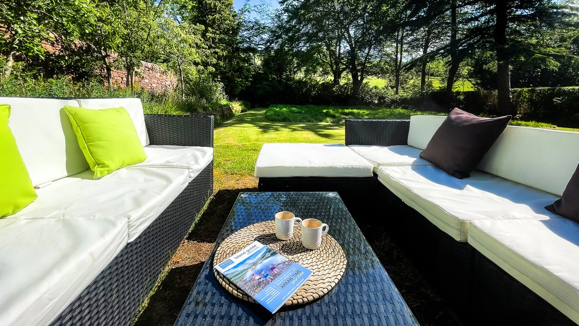 Outdoor patio with black wicker furniture and white cushions, green and black pillows, a glass-top coffee table with two white coffee mugs and a travel guidebook, surrounded by a lush green garden with trees and a grassy lawn.