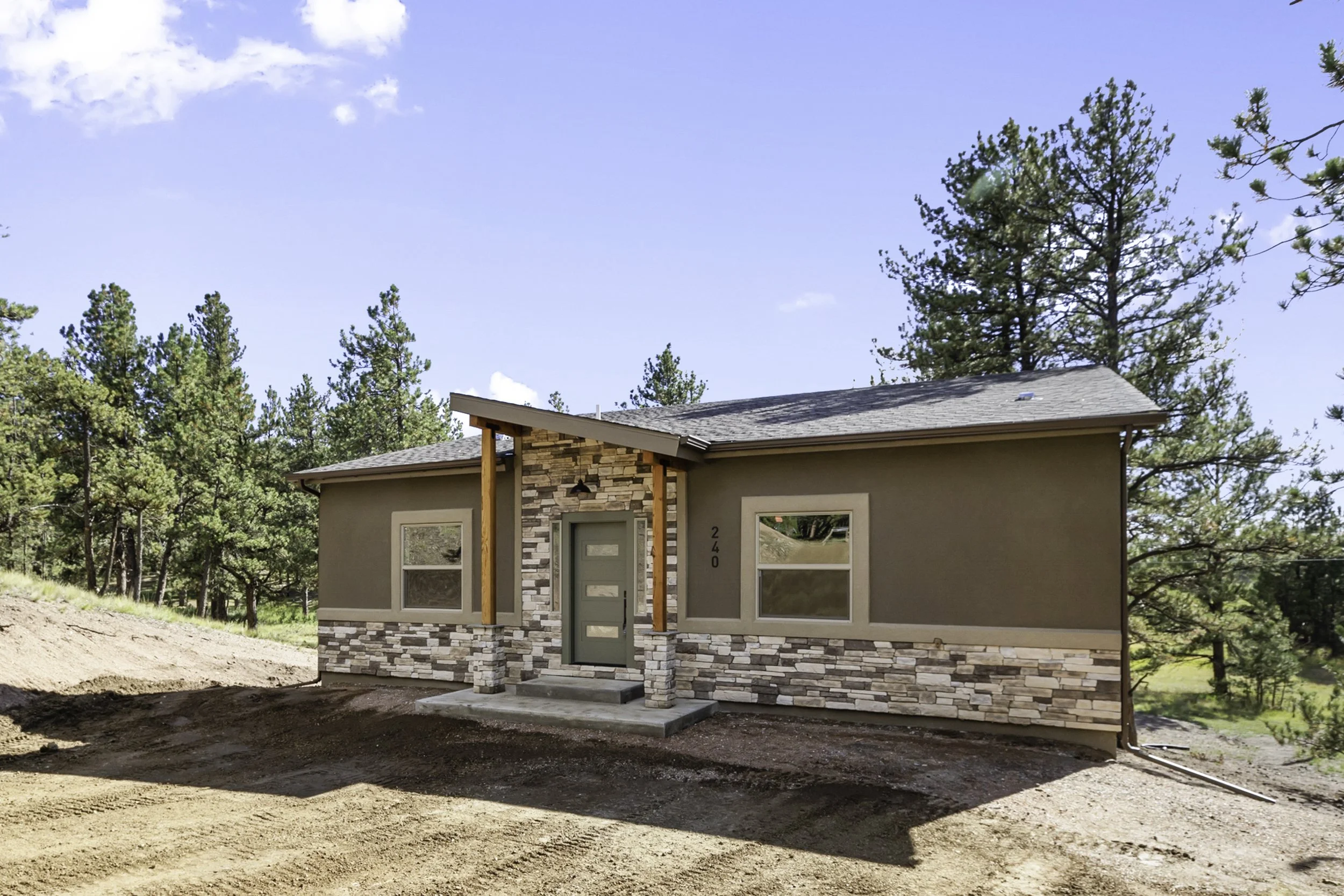 Newly constructed house with tan exterior, large windows, stone accents, and a small front porch, set against a background of tall pine trees under a partly cloudy blue sky.