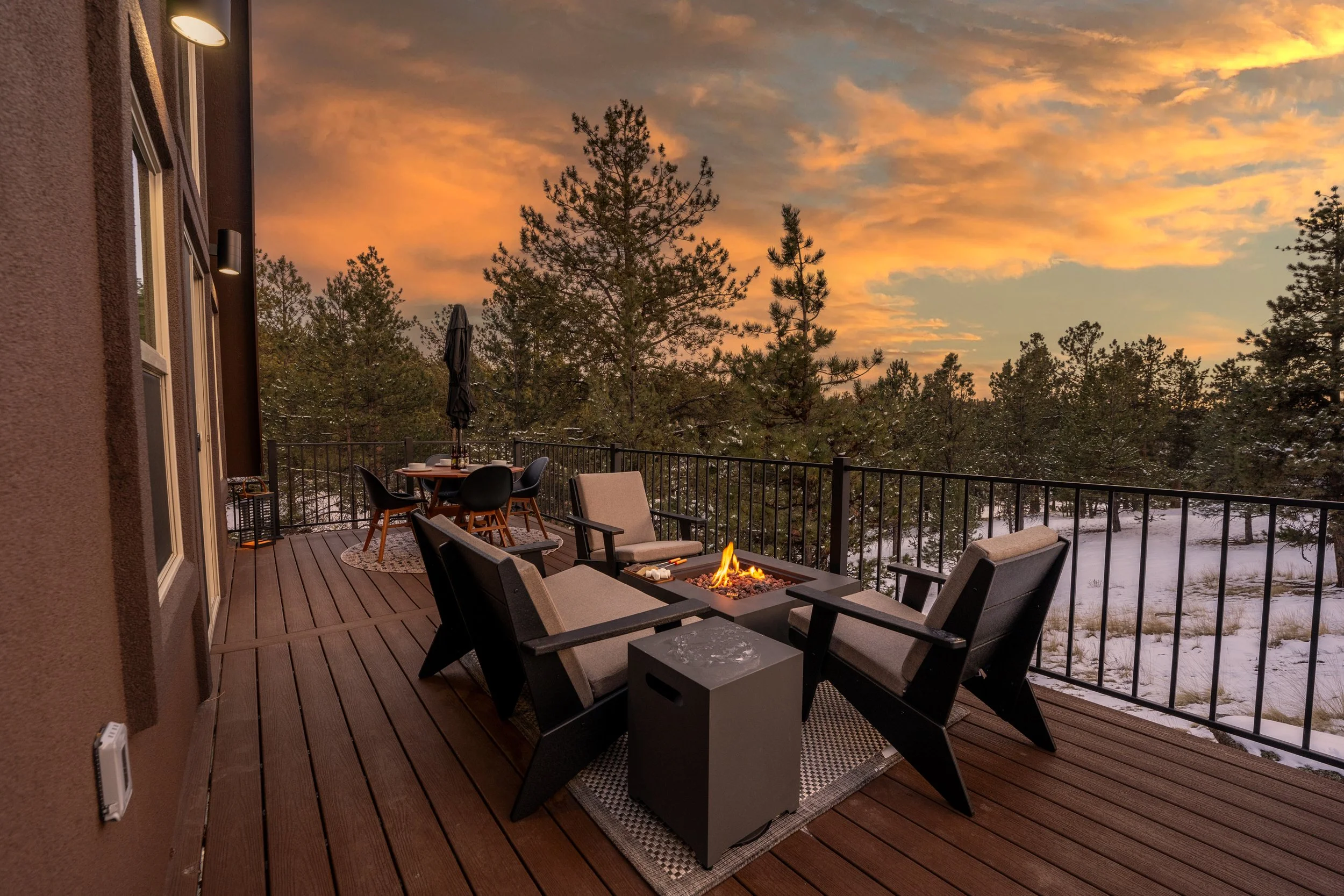 A wooden outdoor balcony with seating and a firepit at sunset, overlooking a wooded landscape with trees and a colorful sky.