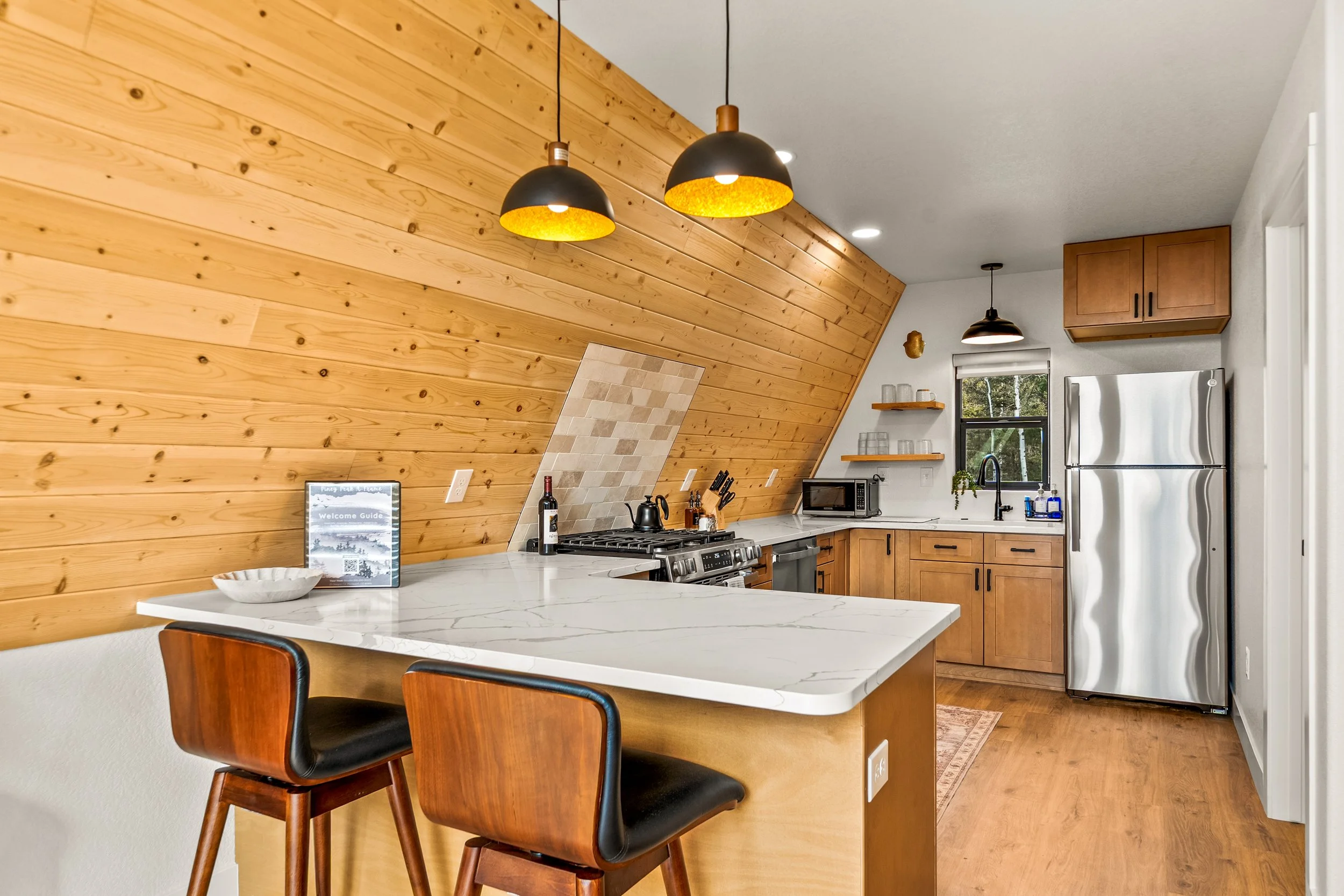 Kitchen with wooden ceiling and cabinets, stainless steel refrigerator, marble countertops, two black and gold pendant lights, window, microwave, and stove.